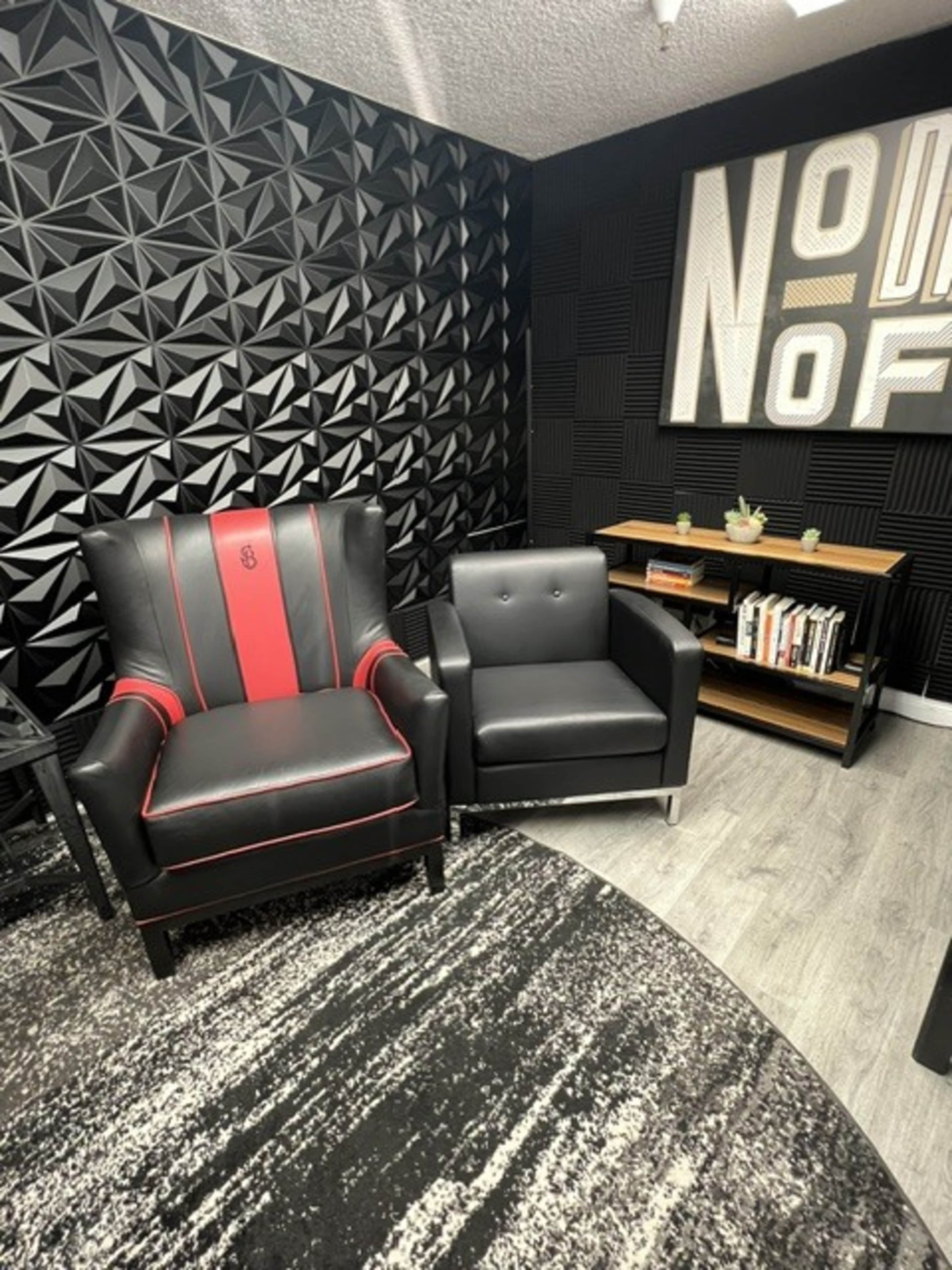 The image shows a modern interior with two distinct chairs, one with red and black upholstery and the other in solid black, set against a textured black wall and accompanied by a wooden shelf filled with books.