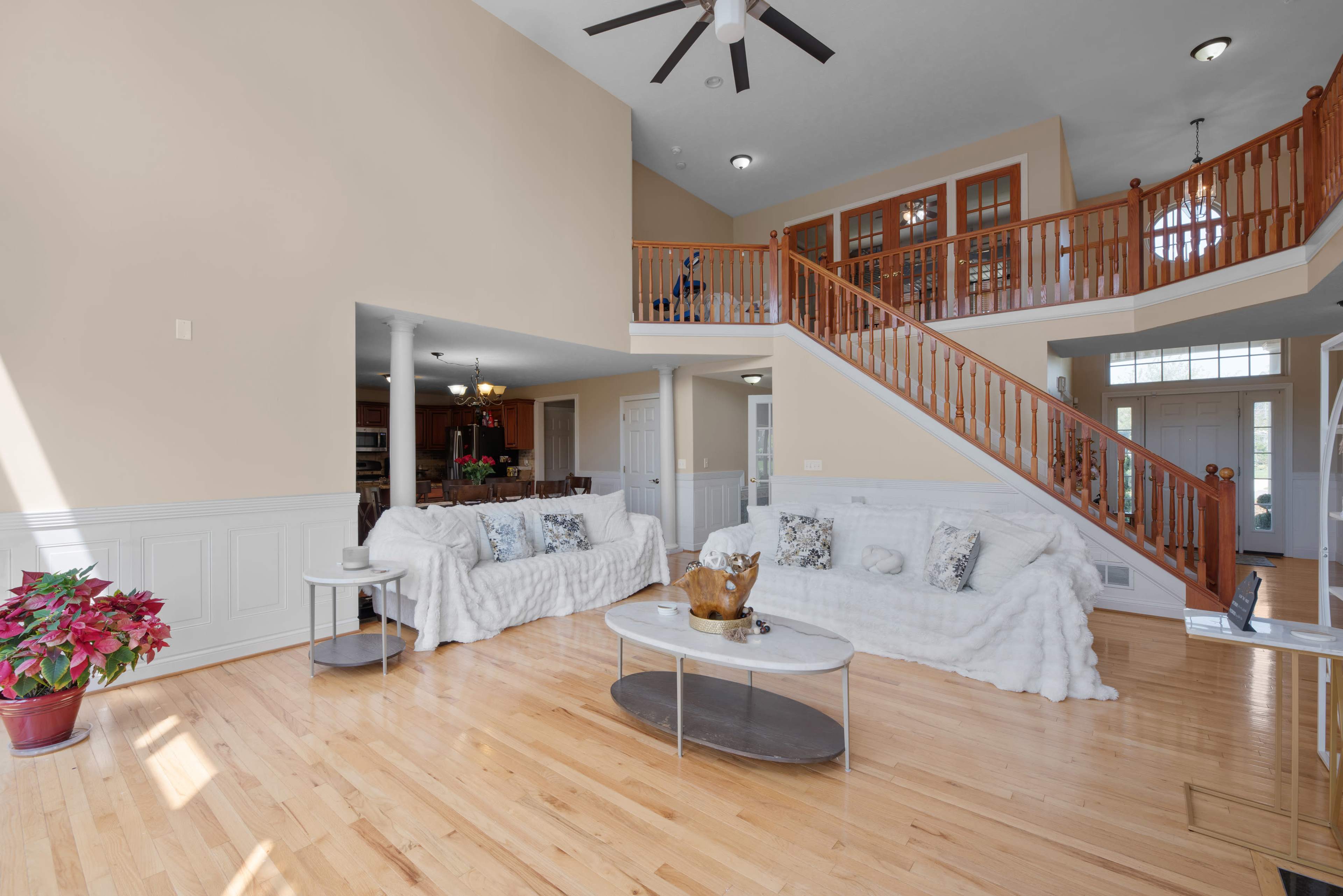 The image depicts a spacious living room with white sofas, a glass coffee table, and a staircase leading to an upper level.