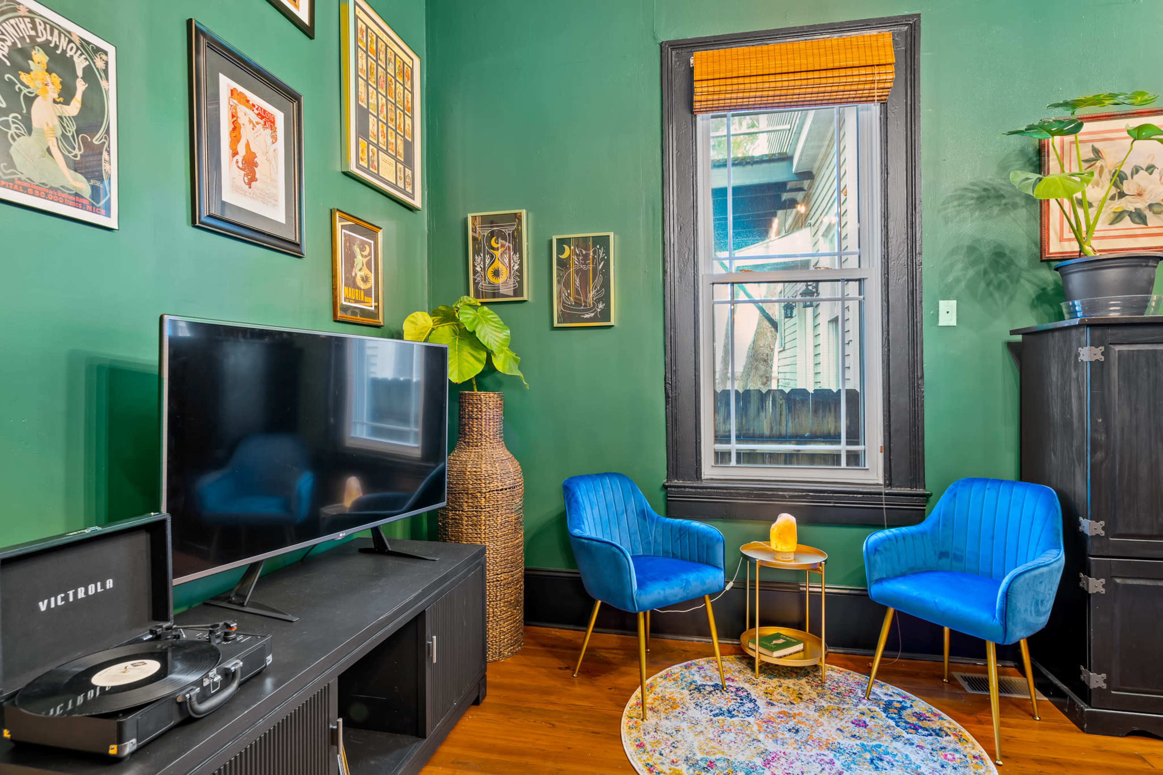 The image features a cozy room with green walls, a television, a record player, two blue chairs, a small golden table, and framed artwork.