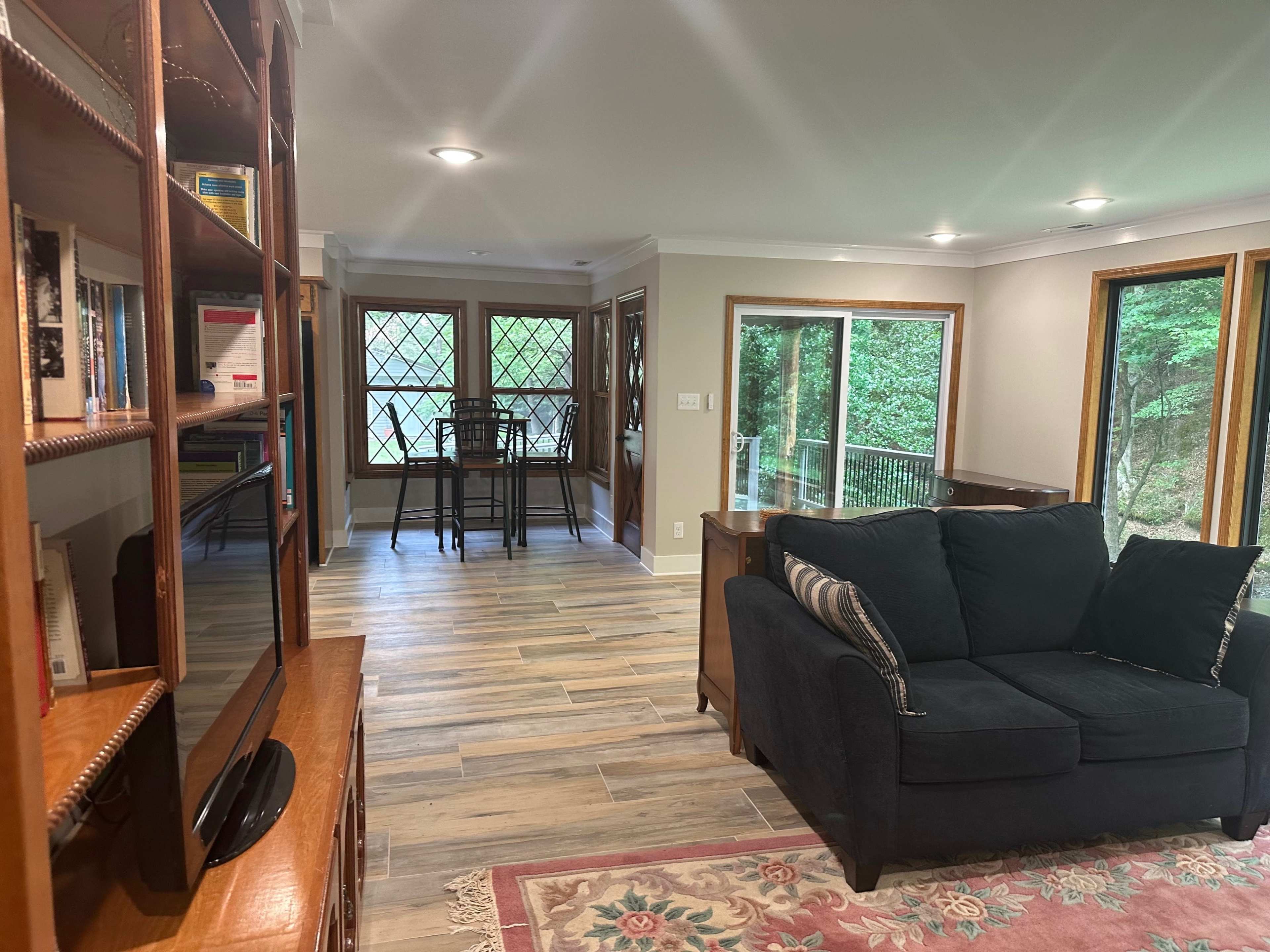 The image shows a living area featuring a dark couch, a wooden bookshelf, and a dining table with chairs visible in the background.