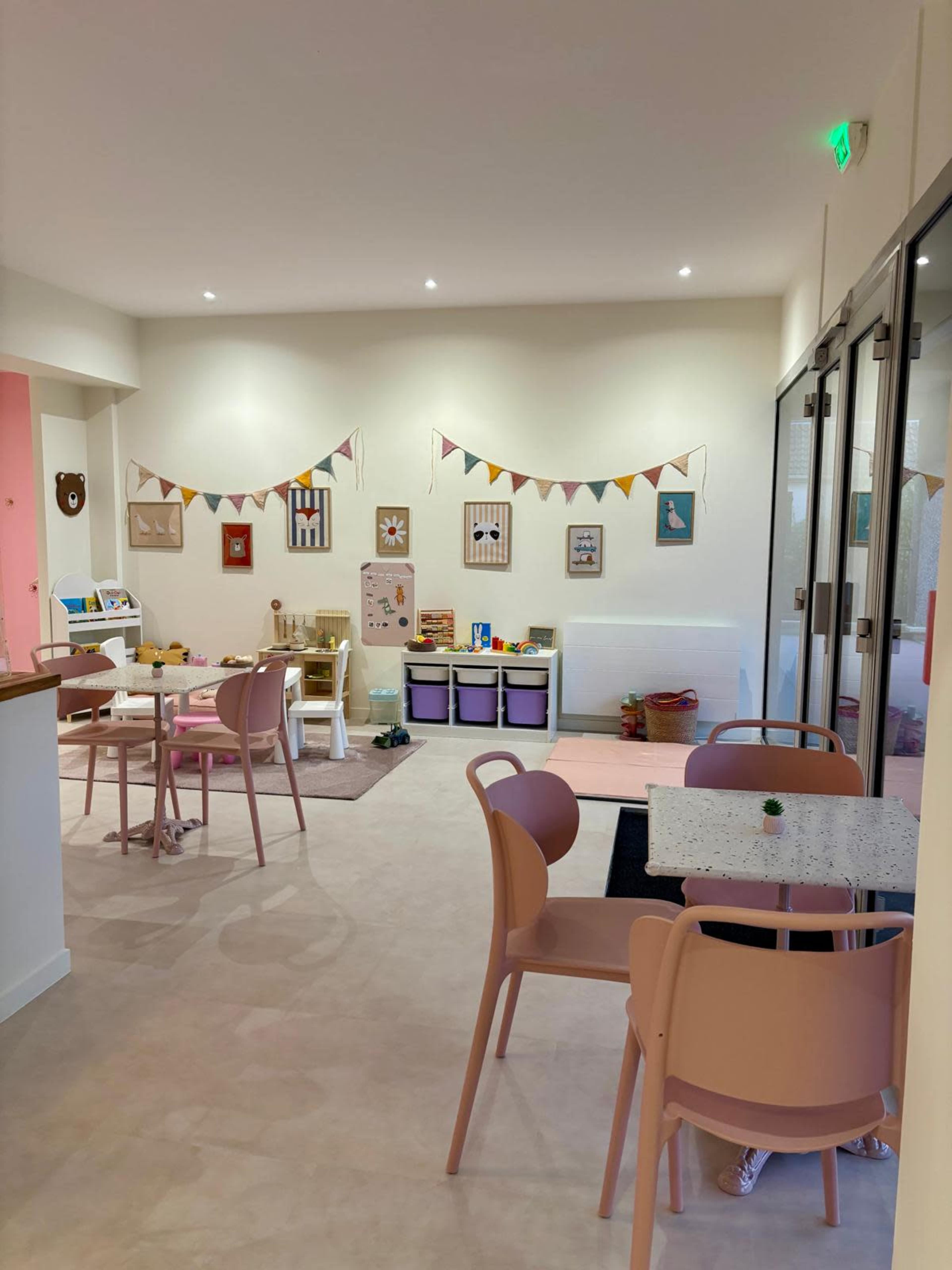 A brightly lit playroom features several tables, colorful decorations, and child-sized furniture, along with a variety of toys and play areas.