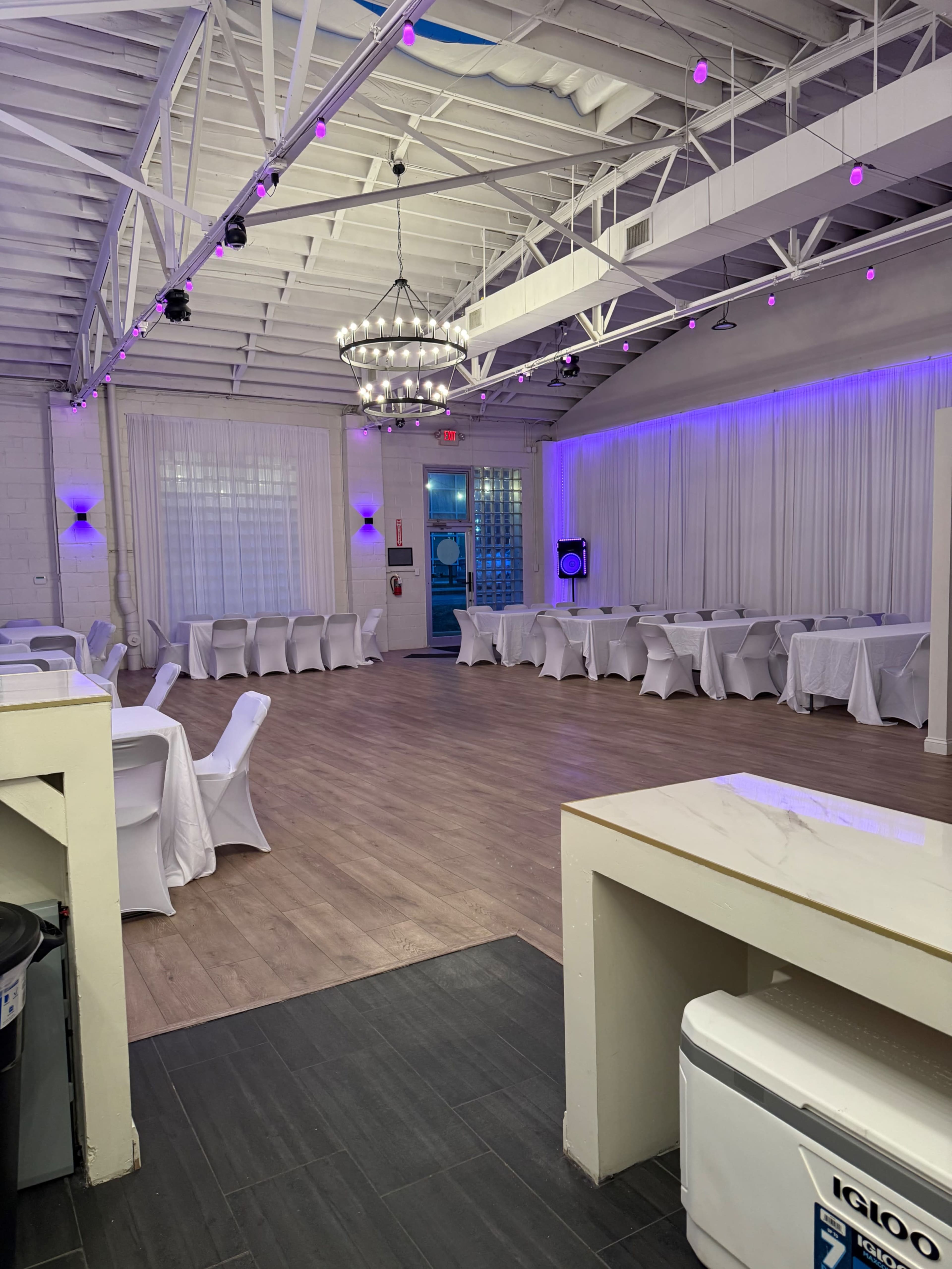 The image shows a spacious event venue with a wooden floor, white draped tables, and purple lighting accents.