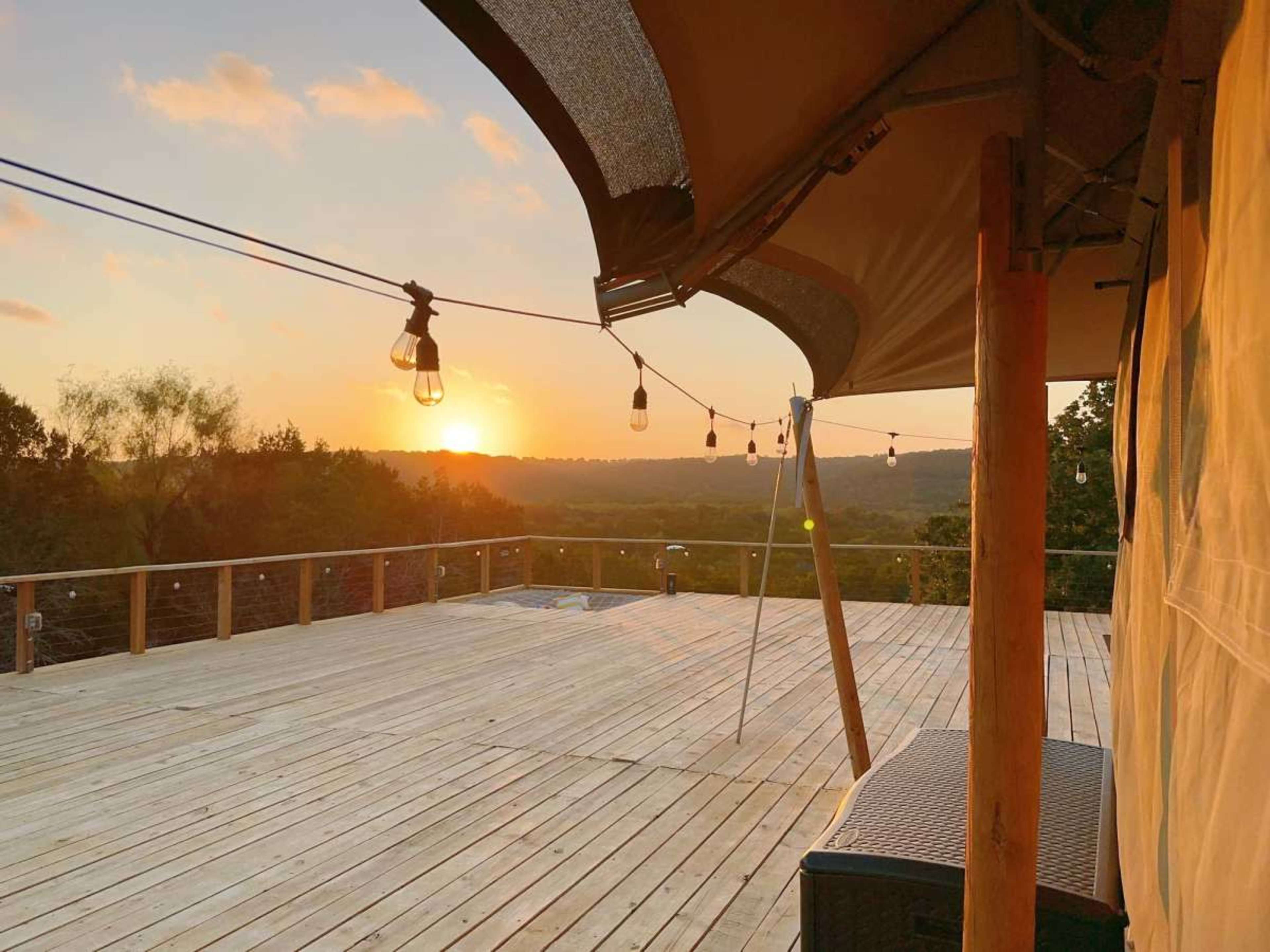 The image shows a wooden deck with a canopy and string lights, overlooking a sunset in a natural landscape.