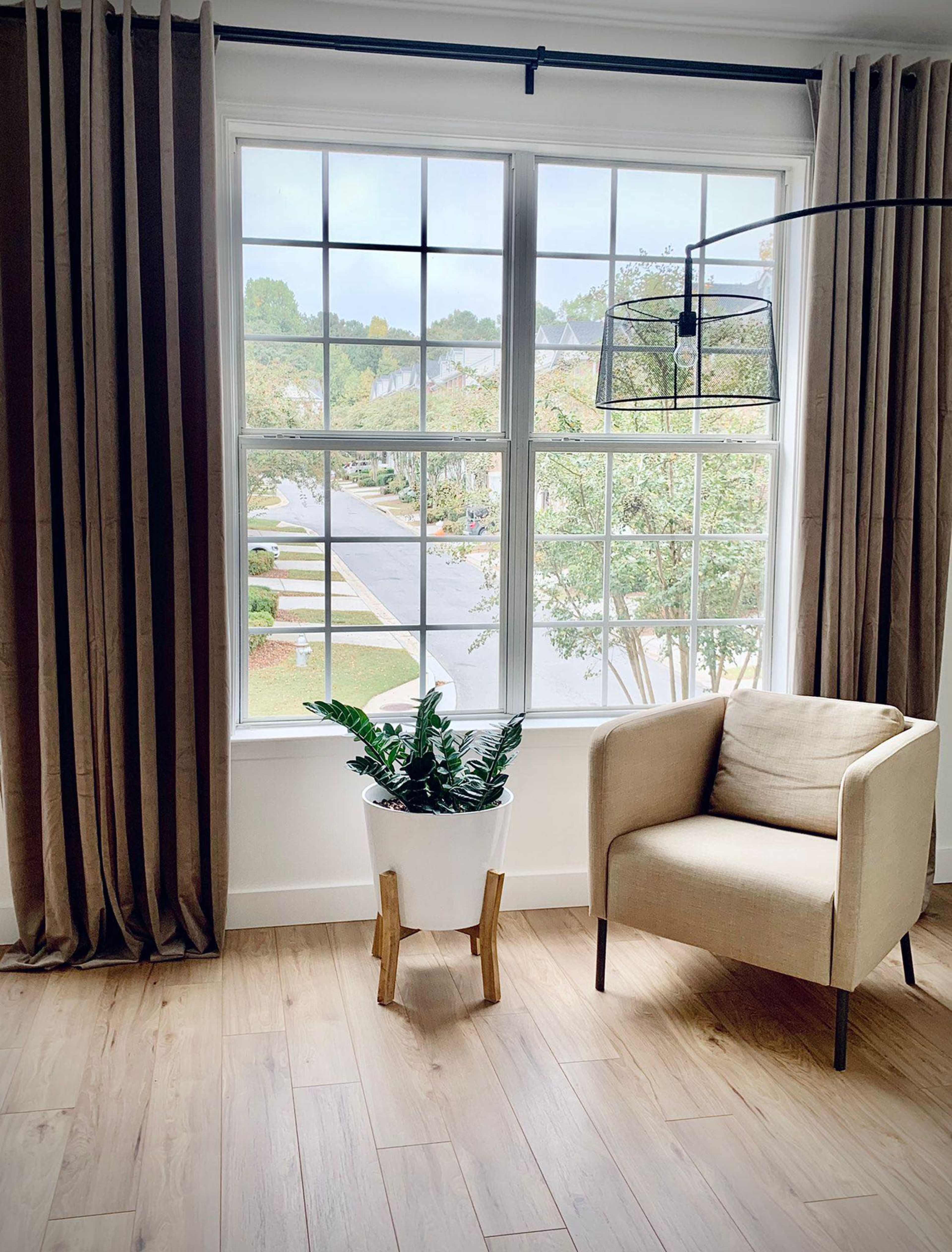 A beige armchair sits beside a large window with a potted plant on the floor beneath it, framed by long curtains.