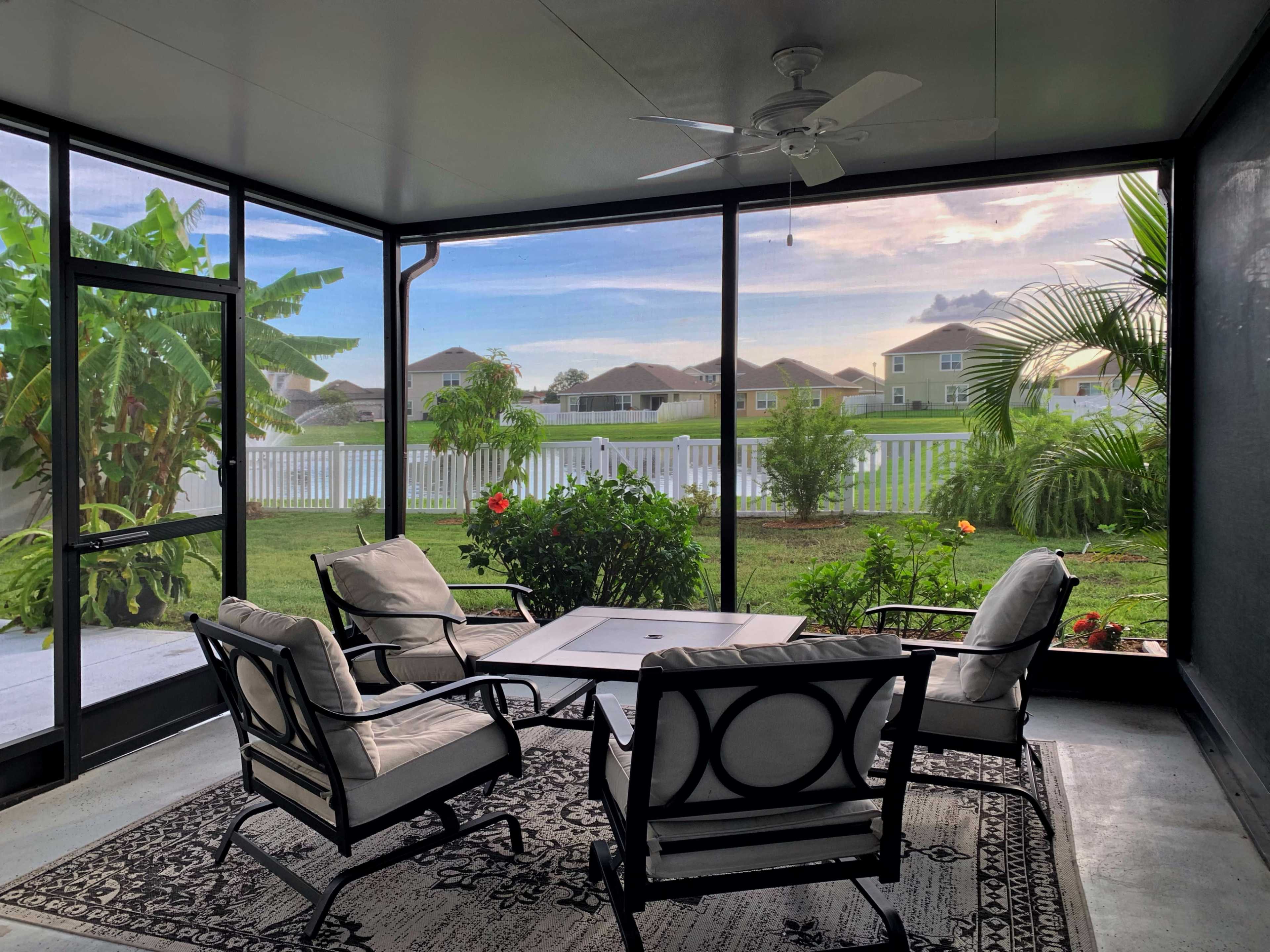 A screened-in porch features four chairs arranged around a coffee table, with a view of a grassy yard and houses in the background.
