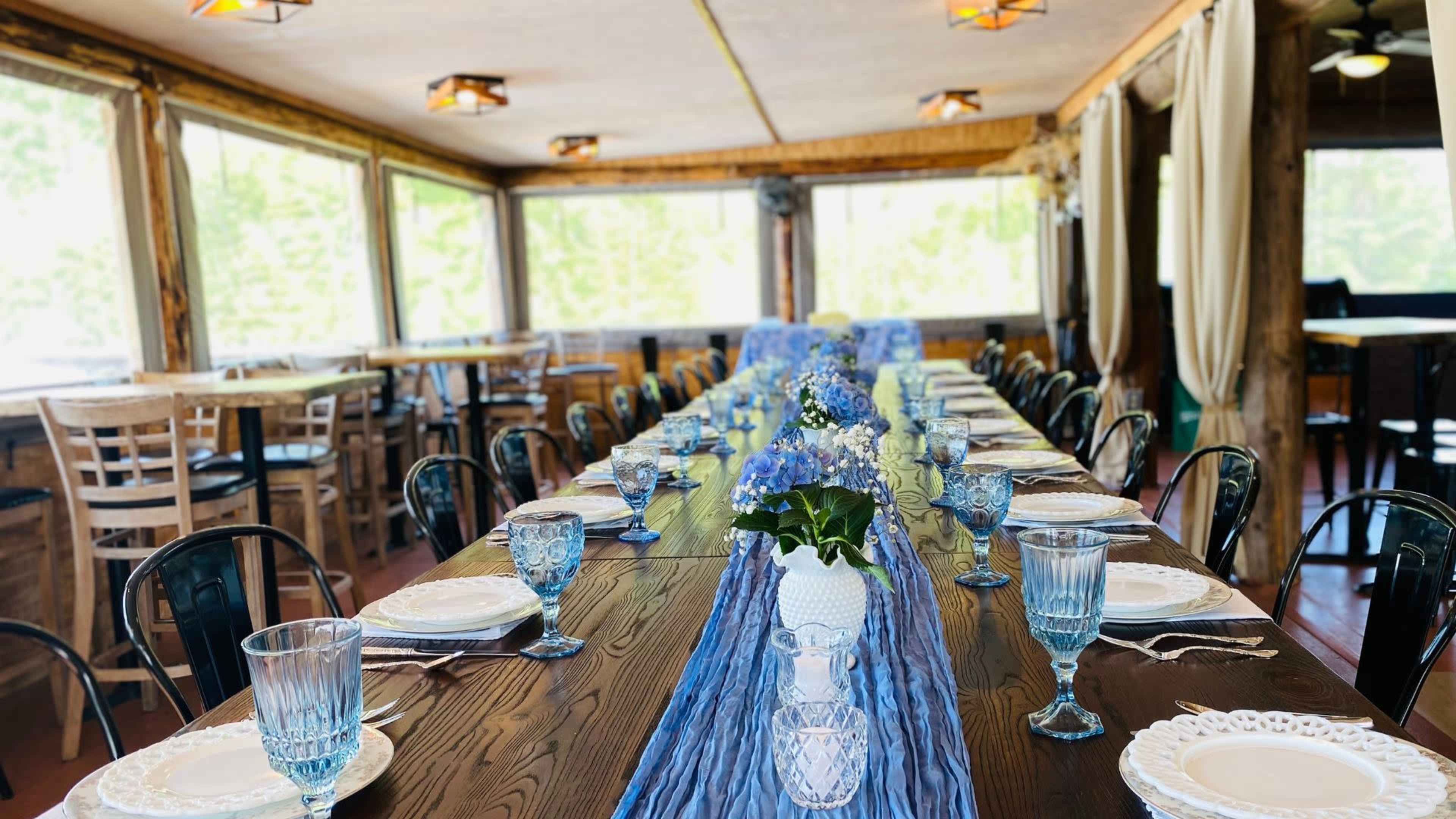 A long dining table set with blue and white dinnerware, decorated with a floral centerpiece and arranged in a rustic indoor dining space with large windows.