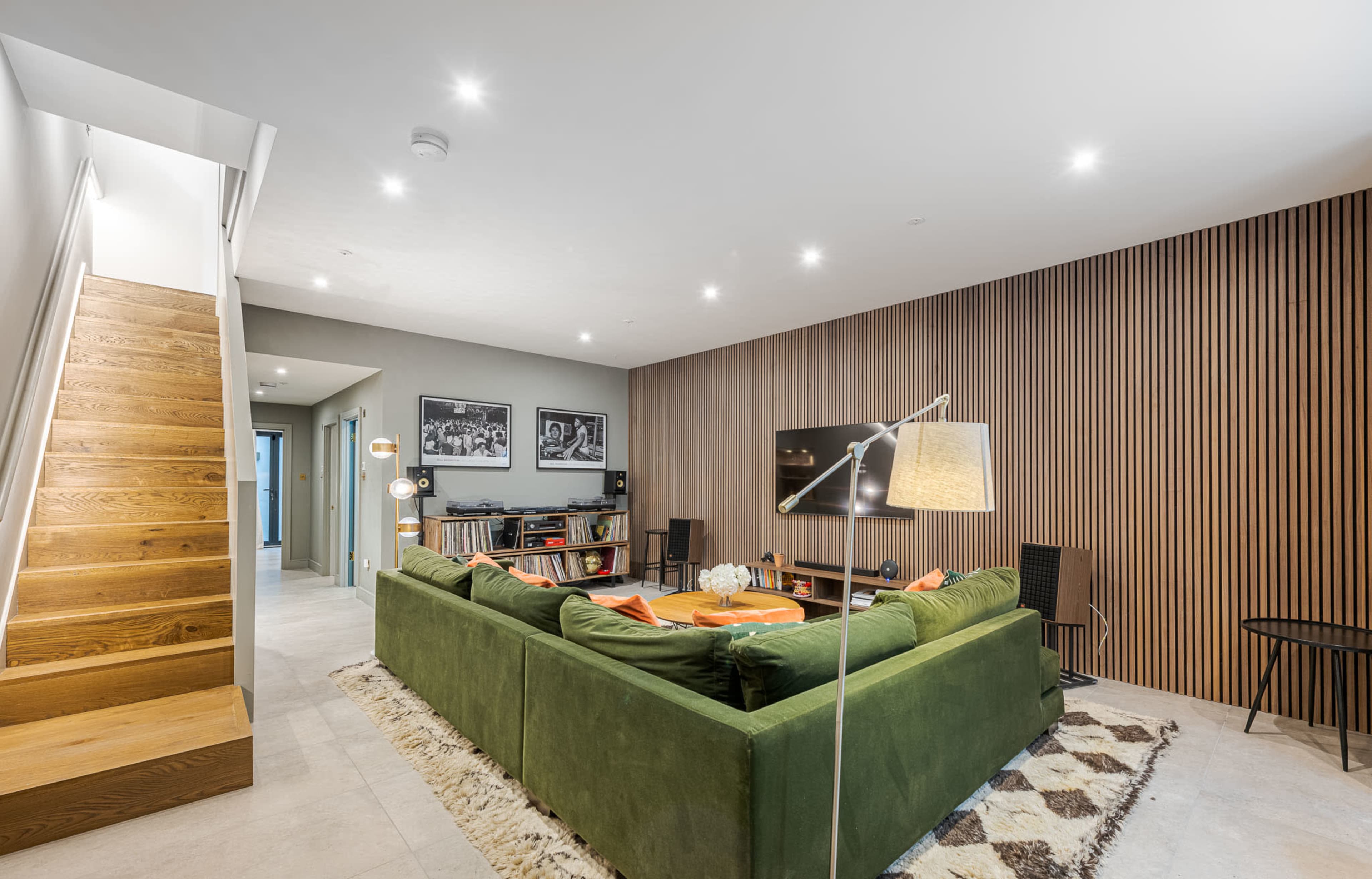 A modern living room features a green sectional sofa, a wooden accent wall, and a staircase leading to the upper level.