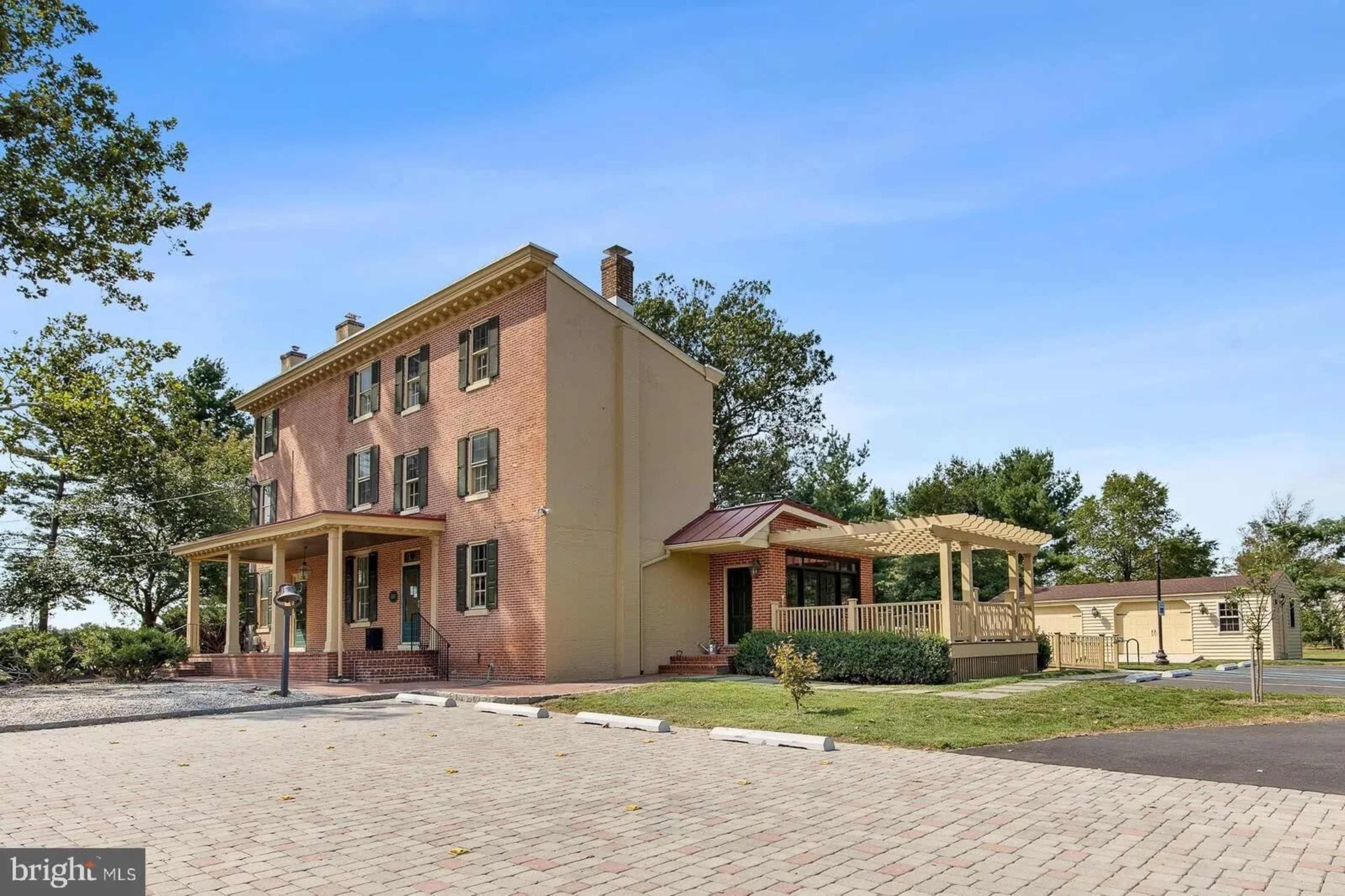 The image shows a two-story brick house with green shutters, a front porch, and a gazebo-style structure beside it, set in a paved area with trees in the background.