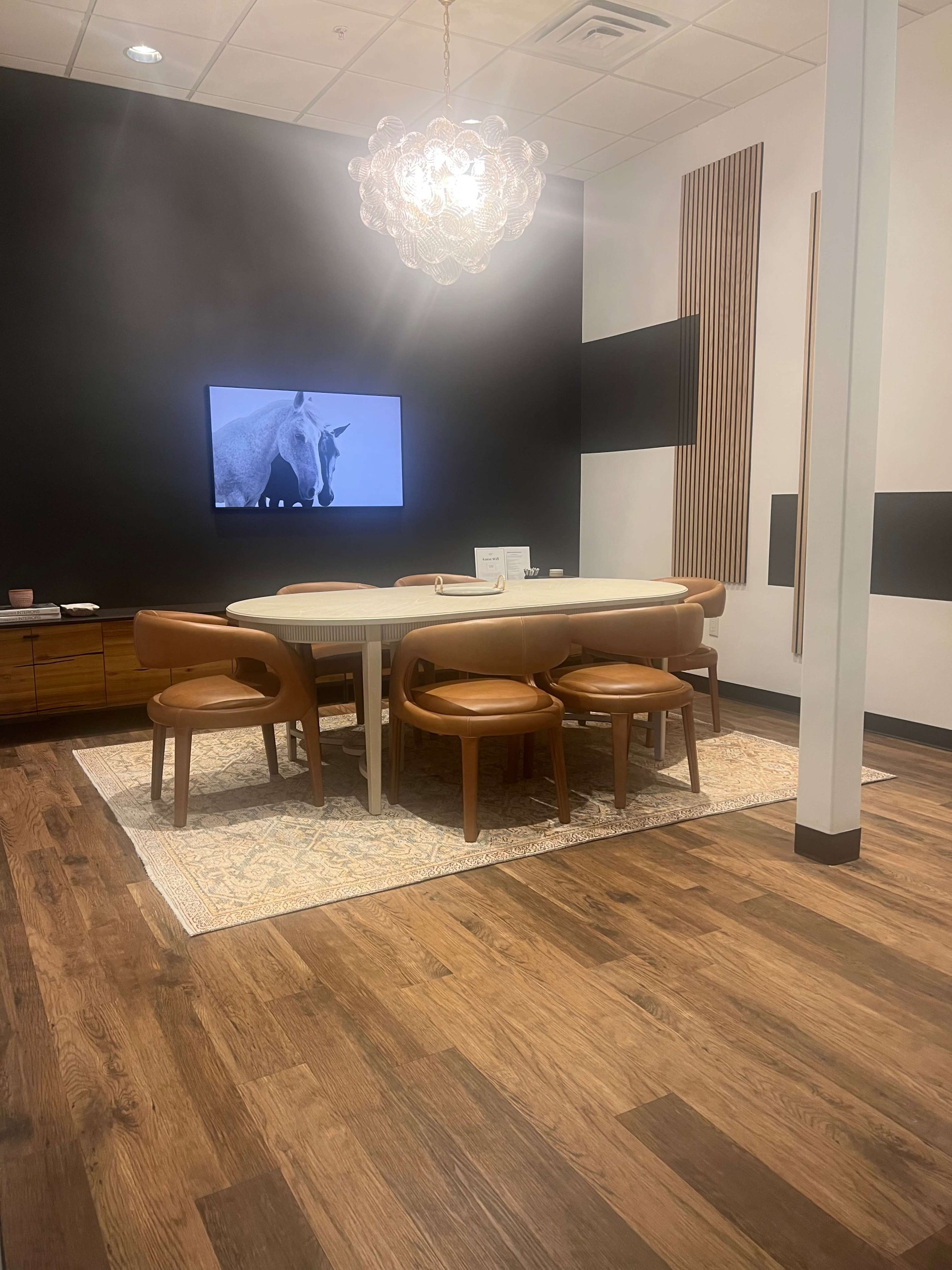 A modern conference room features a rectangular table surrounded by leather chairs, a large screen displaying a horse image, and a pendant light hanging from the ceiling.
