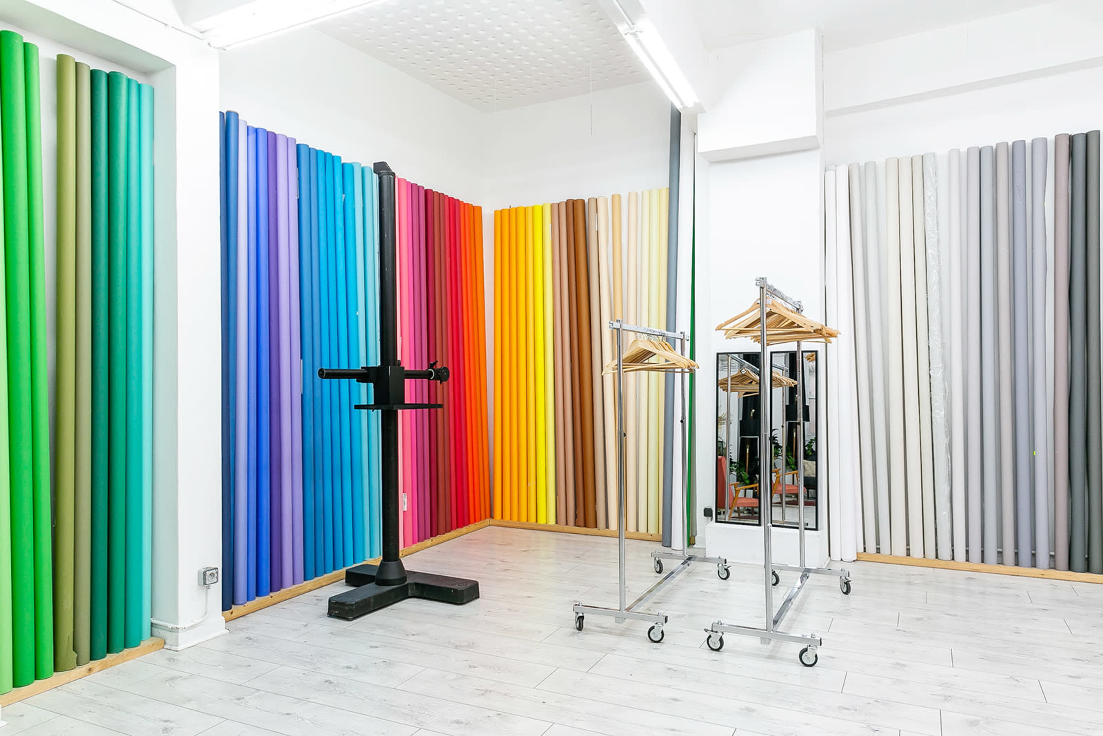 The image shows a spacious studio with colorful backdrop rolls arranged vertically on the walls and clothing racks with hangers positioned in the center.