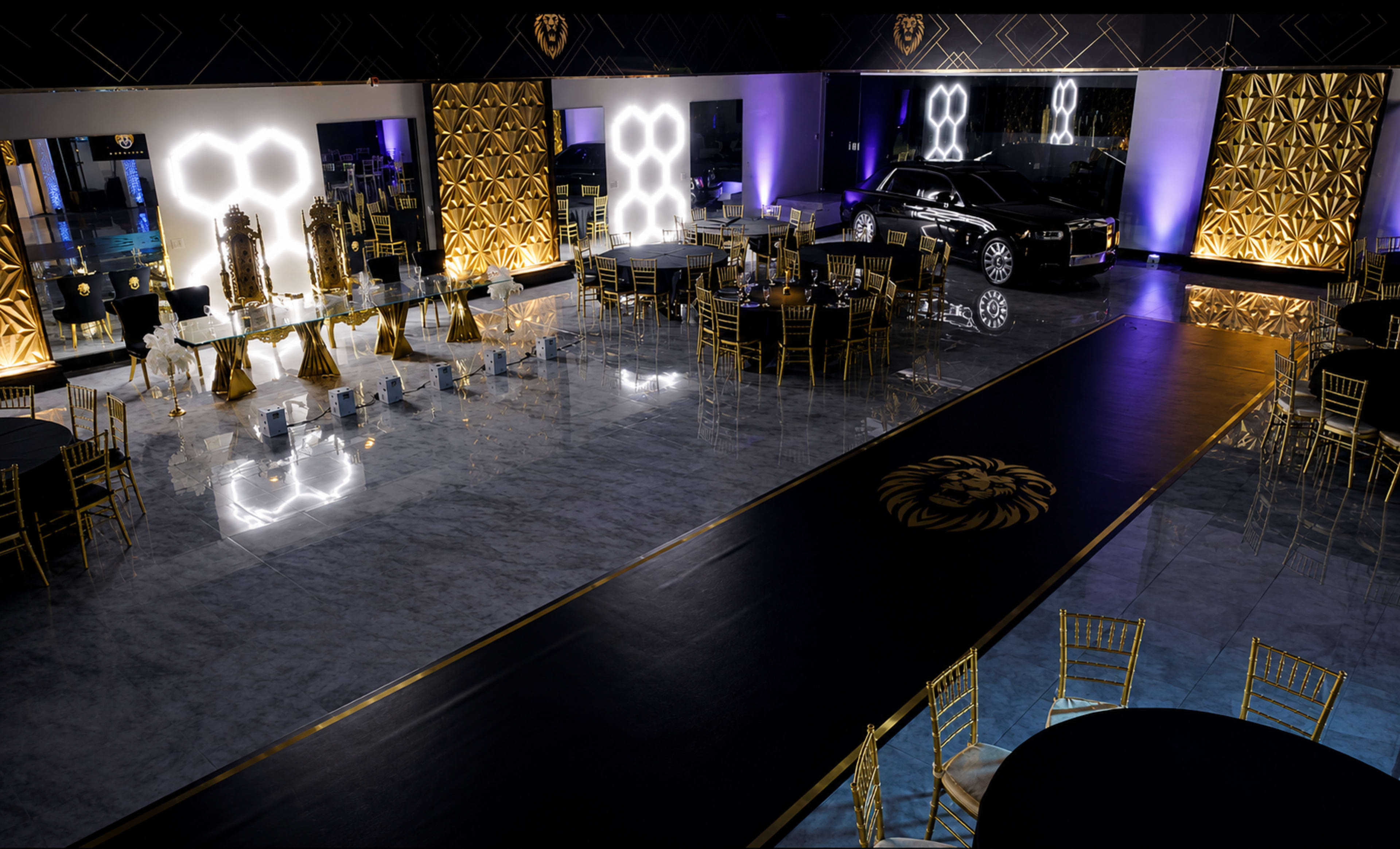 The interior of an elegant event space features a luxurious car, gold-accented tables, and a long runway, all set against a modern backdrop with patterned walls.