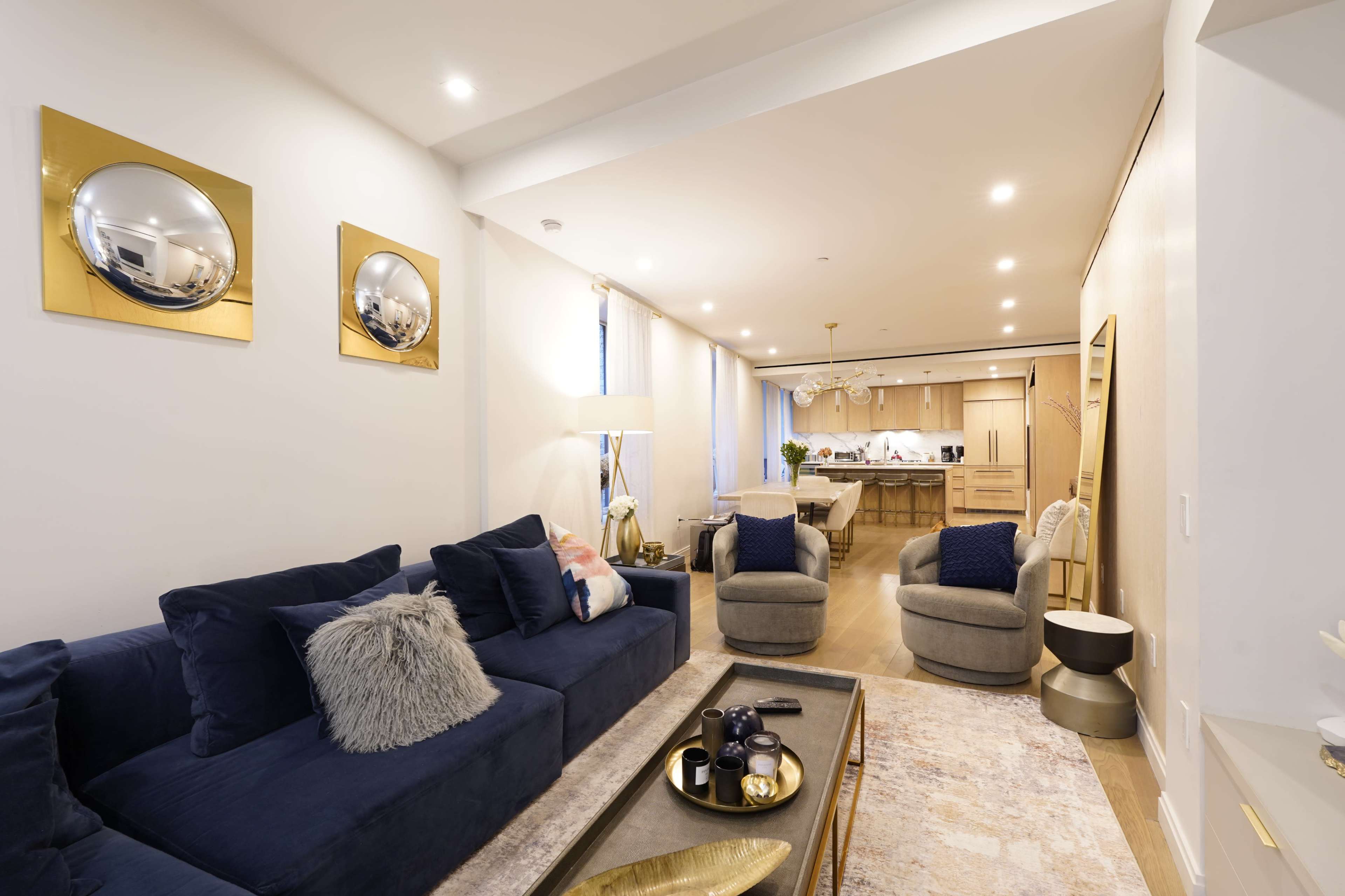 A modern living room features a dark blue sofa, two gray armchairs, and a golden coffee table, adjacent to an open kitchen with light wooden cabinetry.