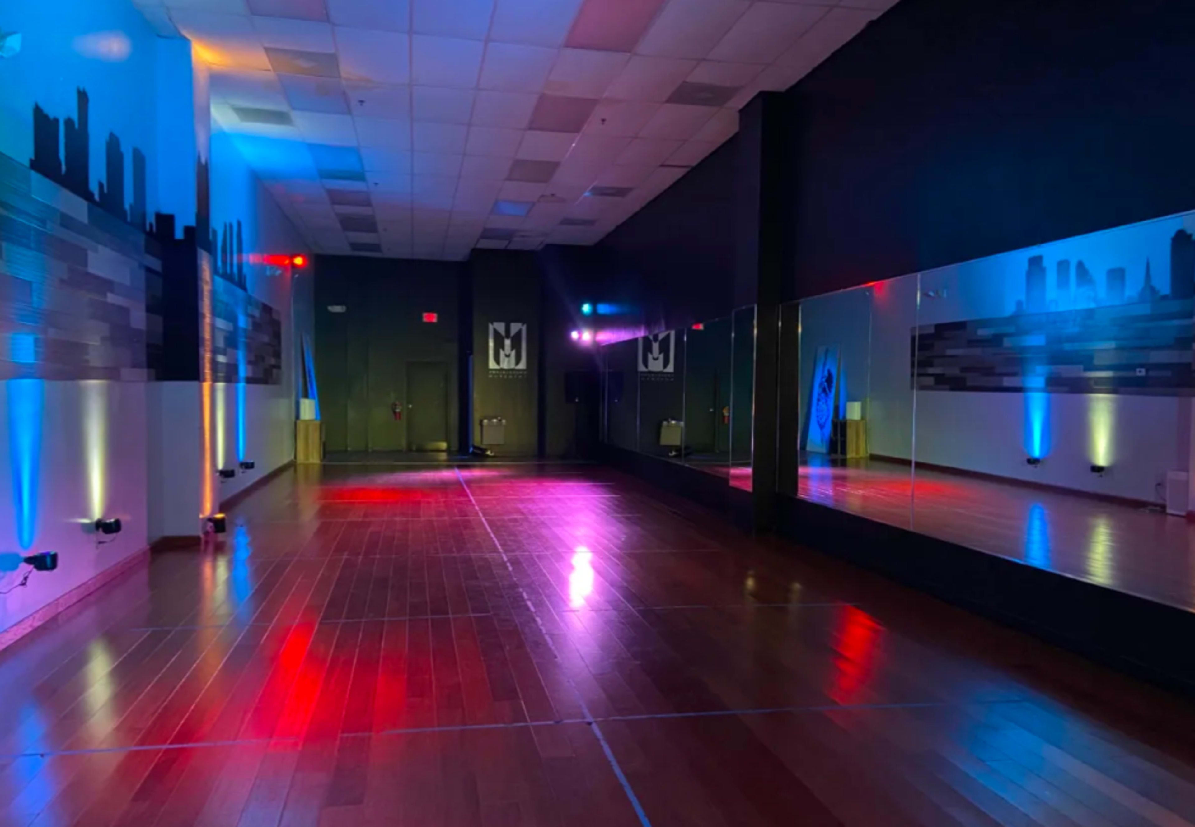 A dance studio with wooden flooring, mirrors on one wall, and colorful lighting along the walls.
