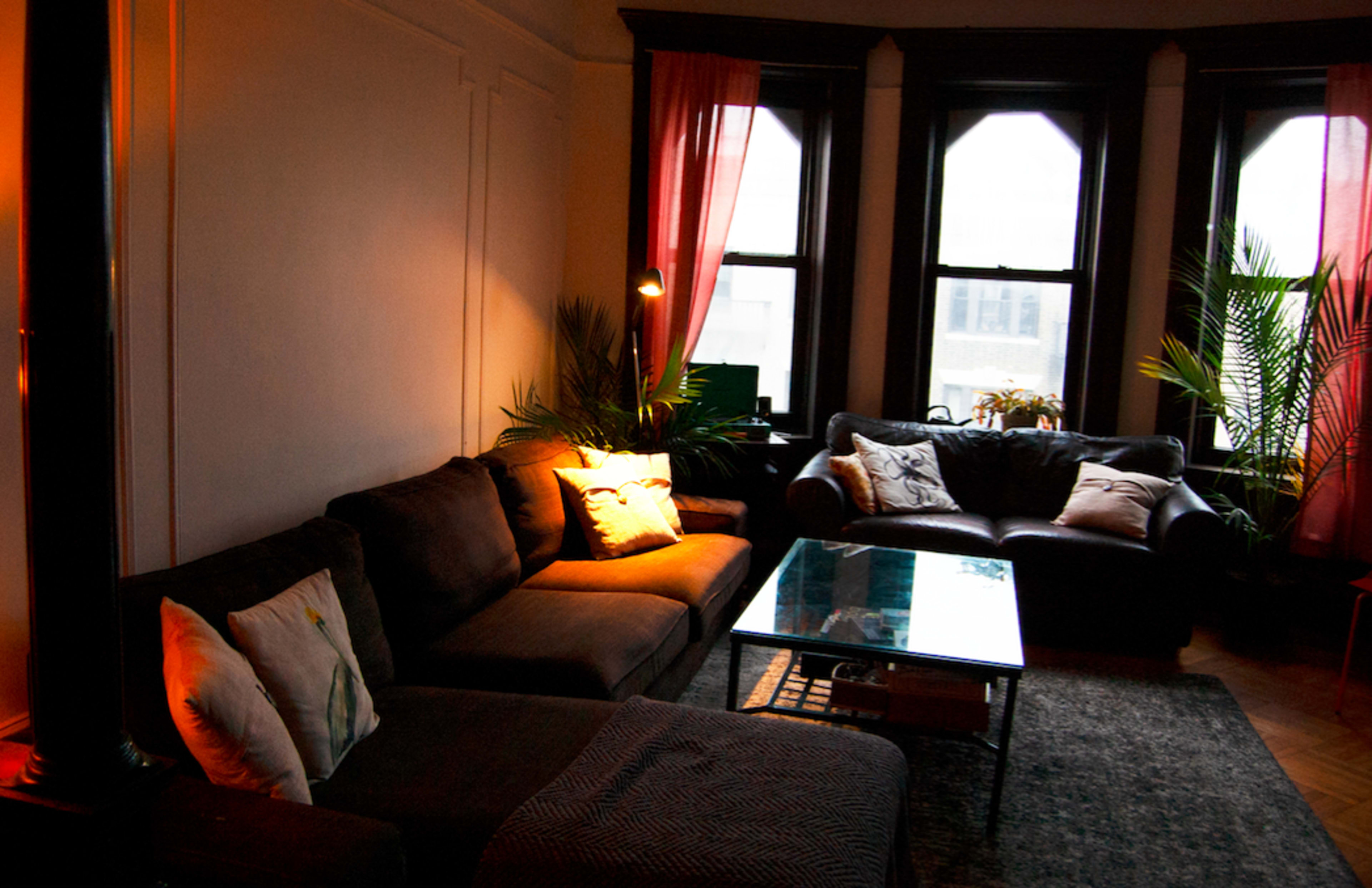 A living room with two sofas, a glass coffee table, and several windows with light curtains, illuminated by a floor lamp.
