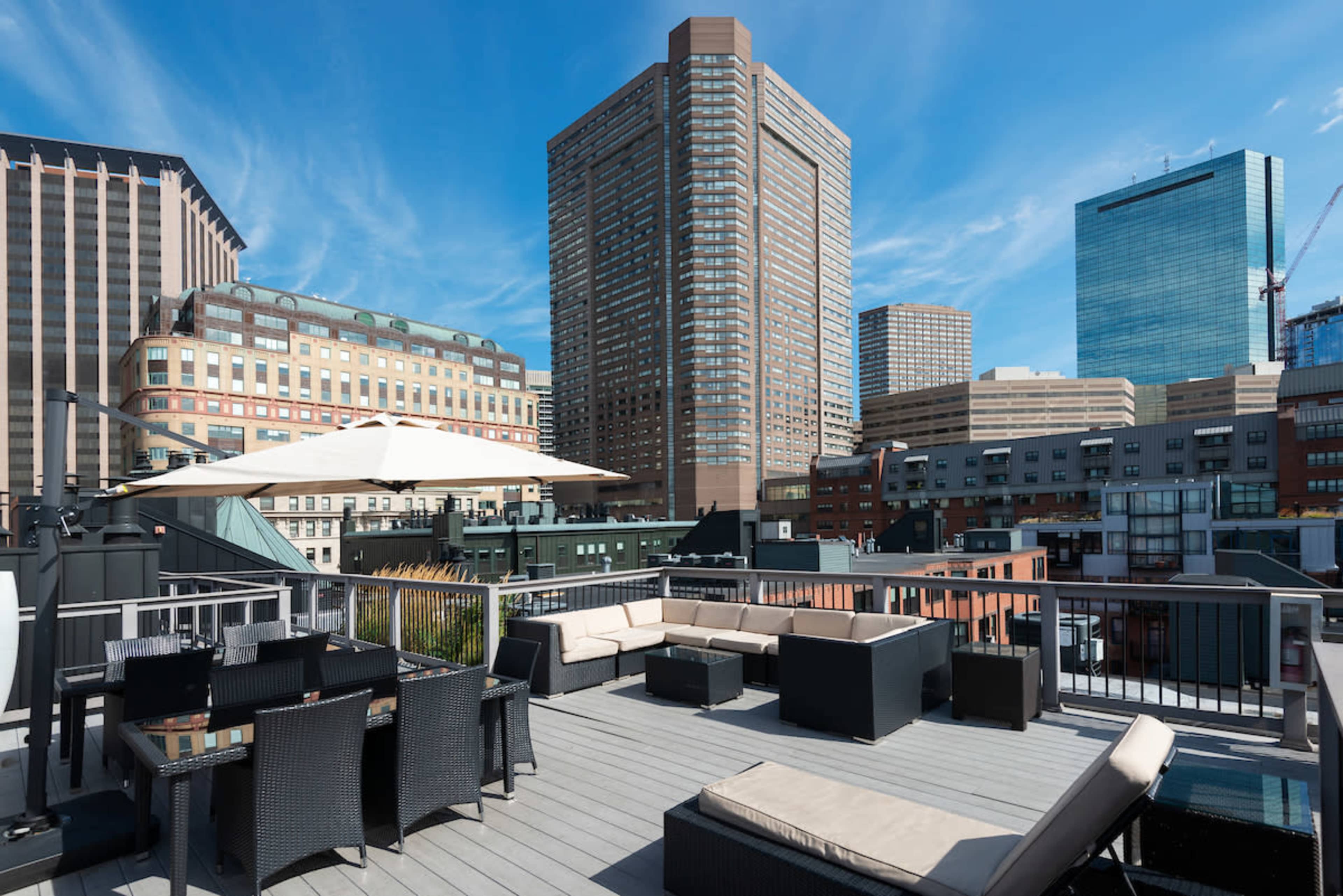 A rooftop terrace features modern outdoor furniture with a view of tall buildings and a clear blue sky.