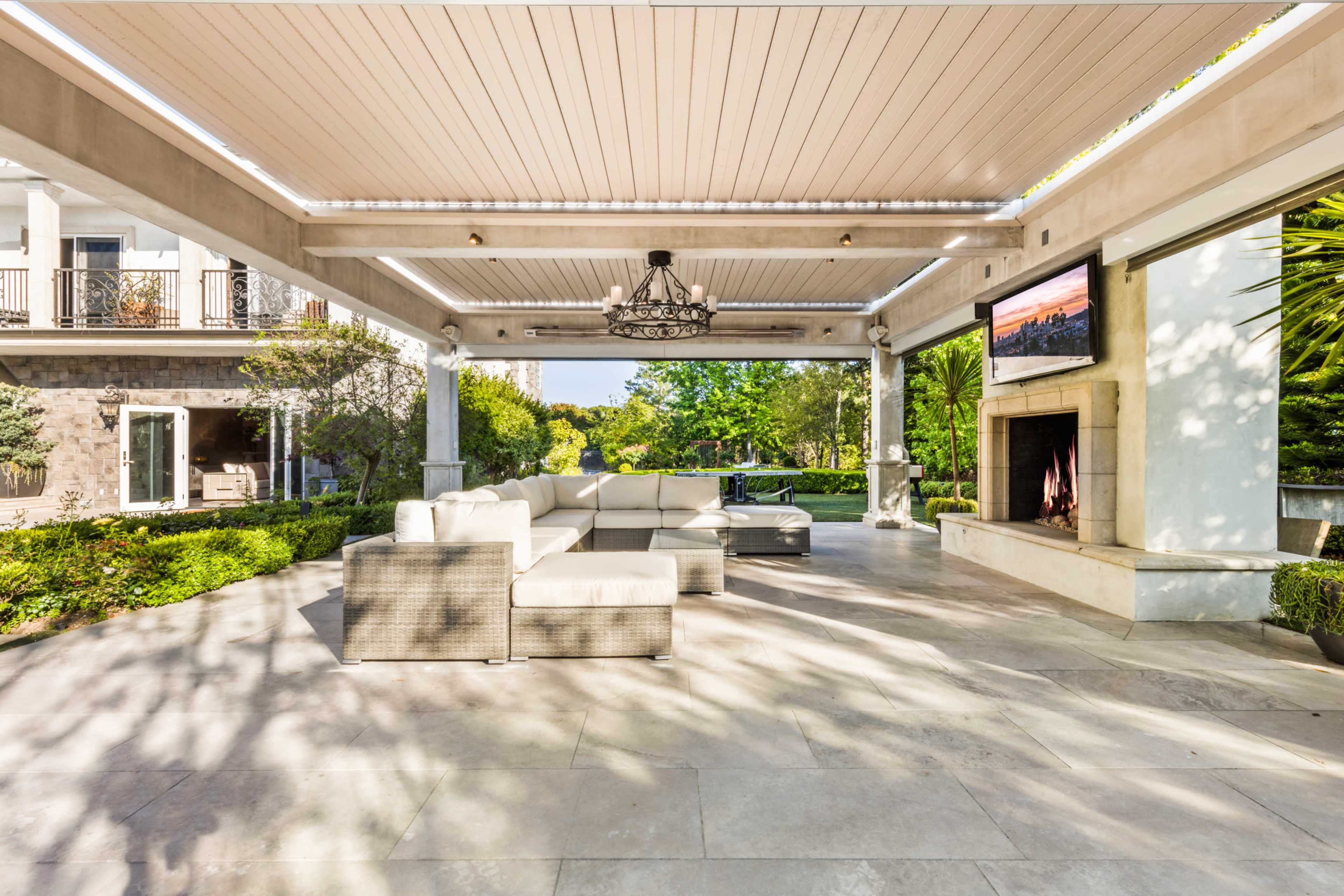 A spacious outdoor patio with a modern sectional sofa, a decorative chandelier, a fireplace, and a mounted television under a covered ceiling.