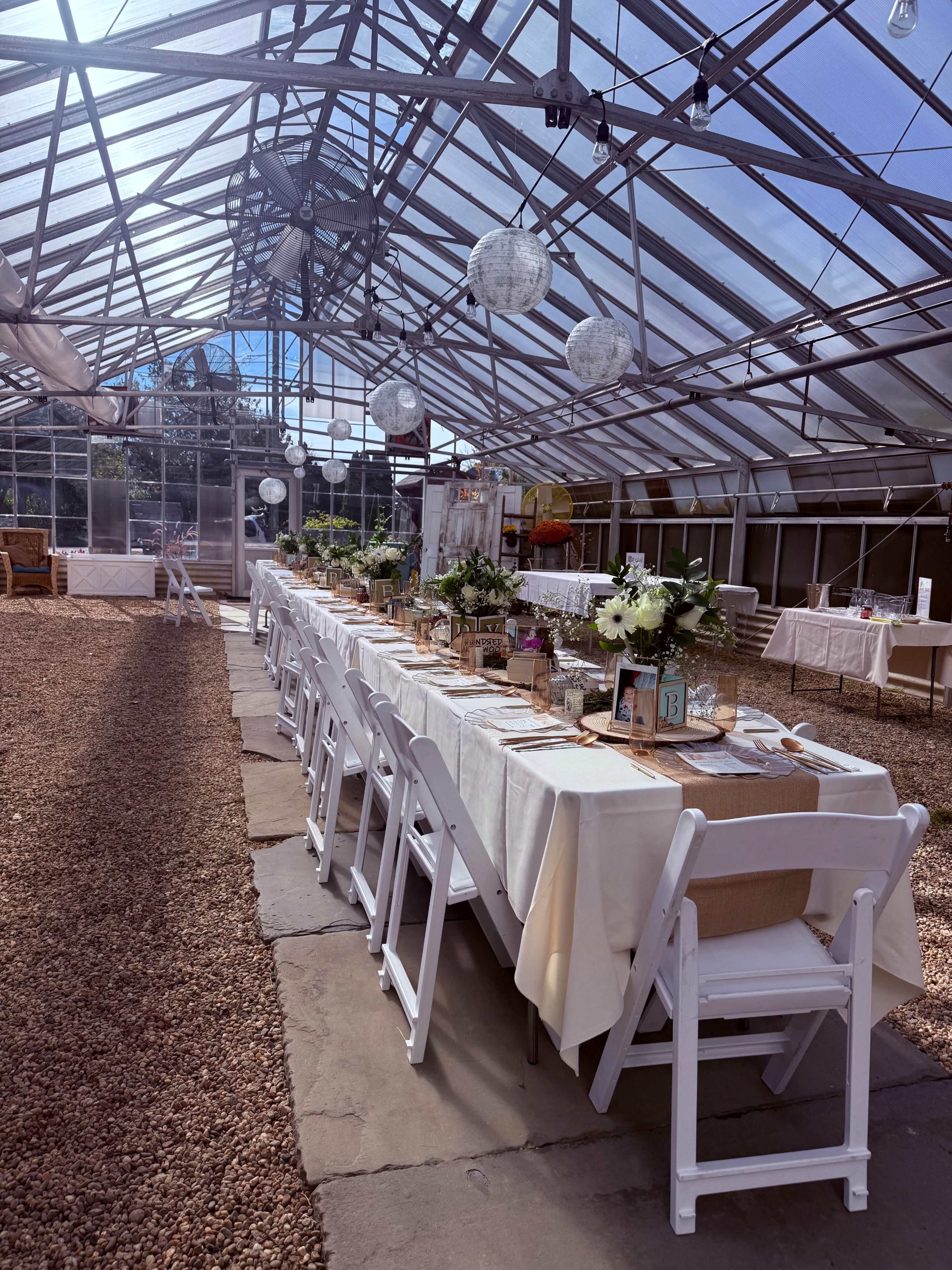 The image shows a long dining table set for an event inside a greenhouse, with floral arrangements, table settings, and decorative lanterns overhead.