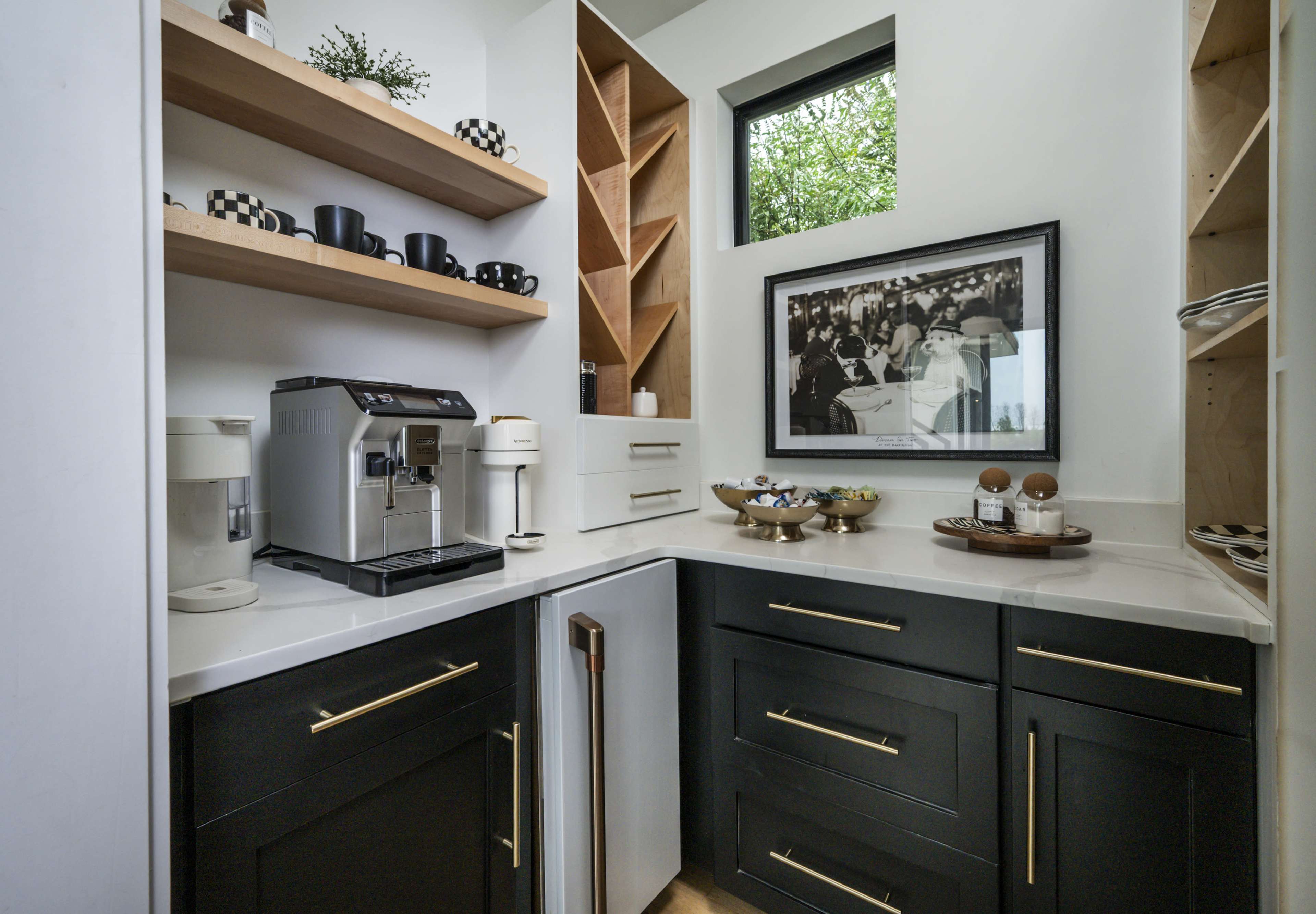 The image shows a modern kitchenette with a coffee machine, wine storage, and shelves displaying cups and a framed black-and-white photograph.