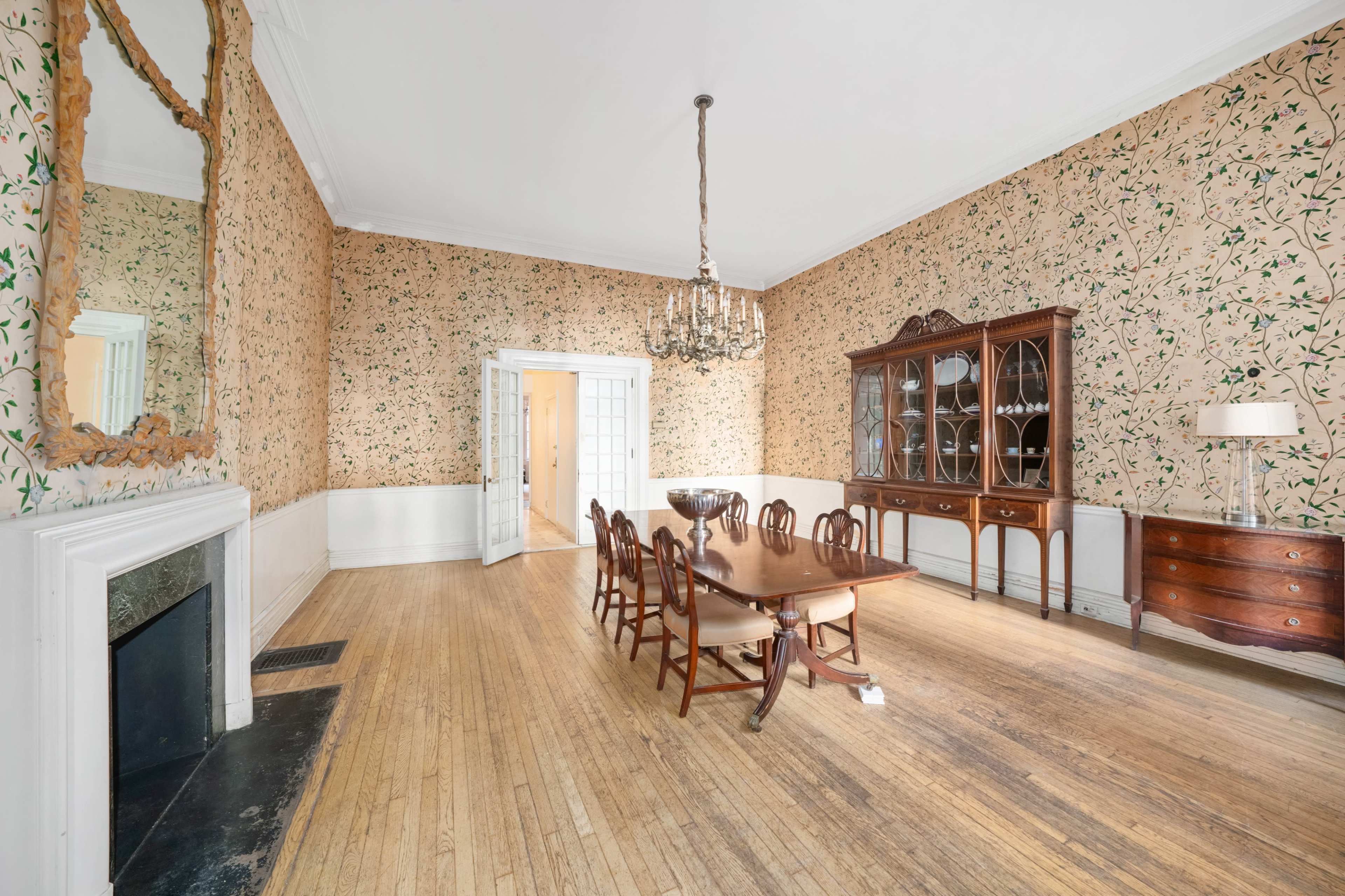 A dining room features a wooden table surrounded by chairs, with floral-patterned wallpaper and a glass-fronted cabinet on one wall.