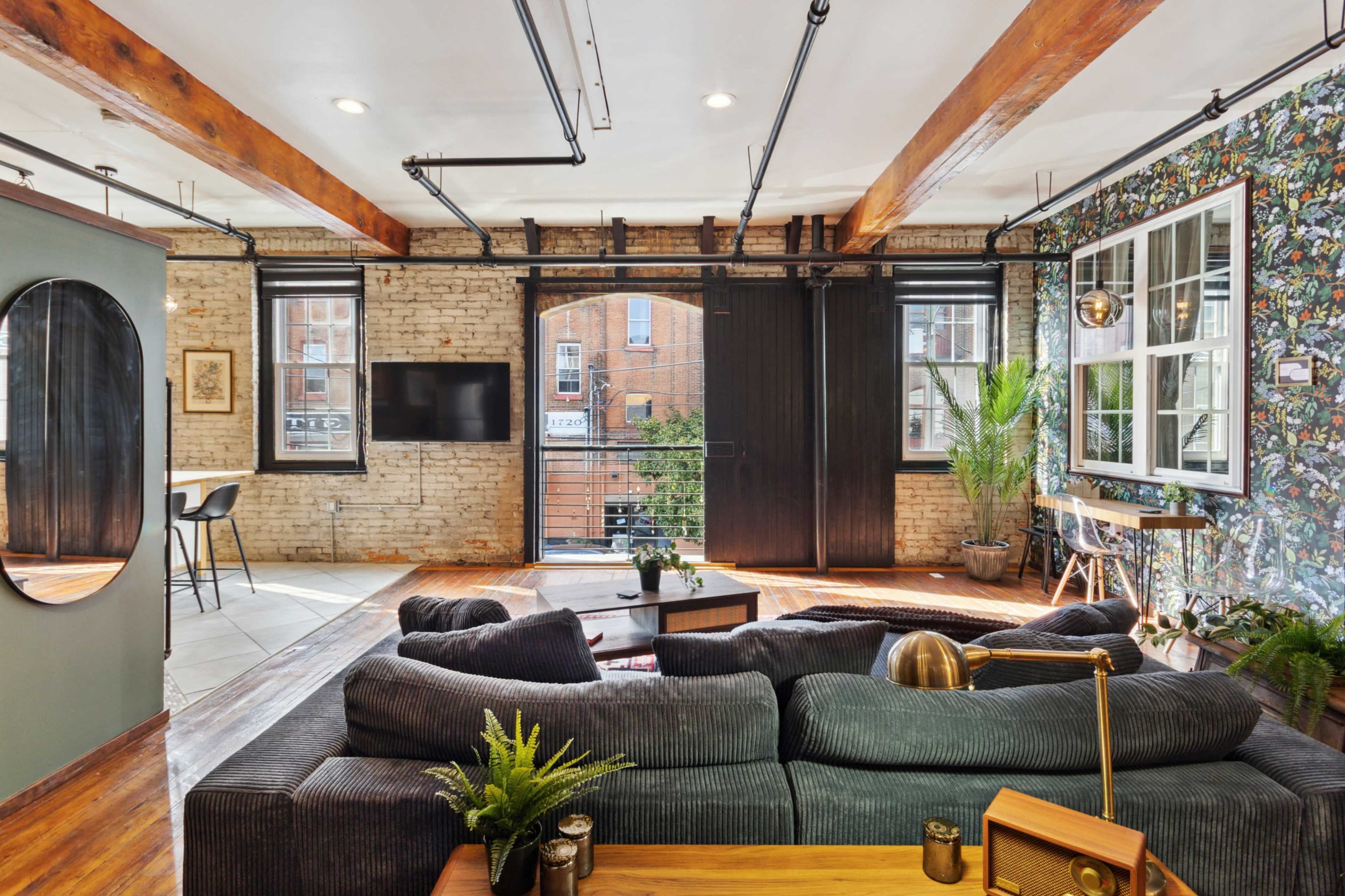A spacious living area featuring exposed brick walls, wooden beams, large windows, and a mix of modern and vintage decor.