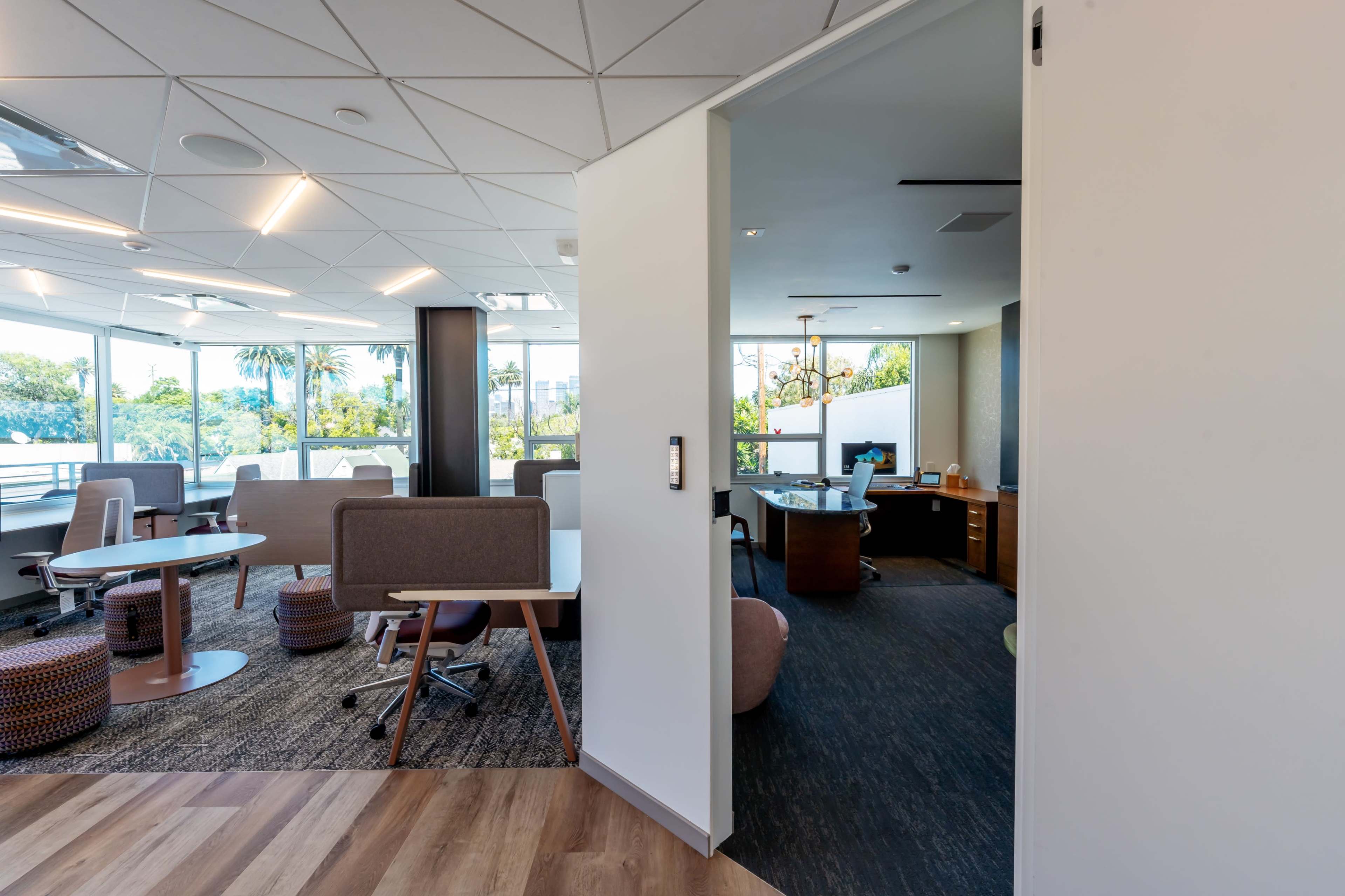 A modern office space with a view of palm trees, featuring various seating areas and workstations.