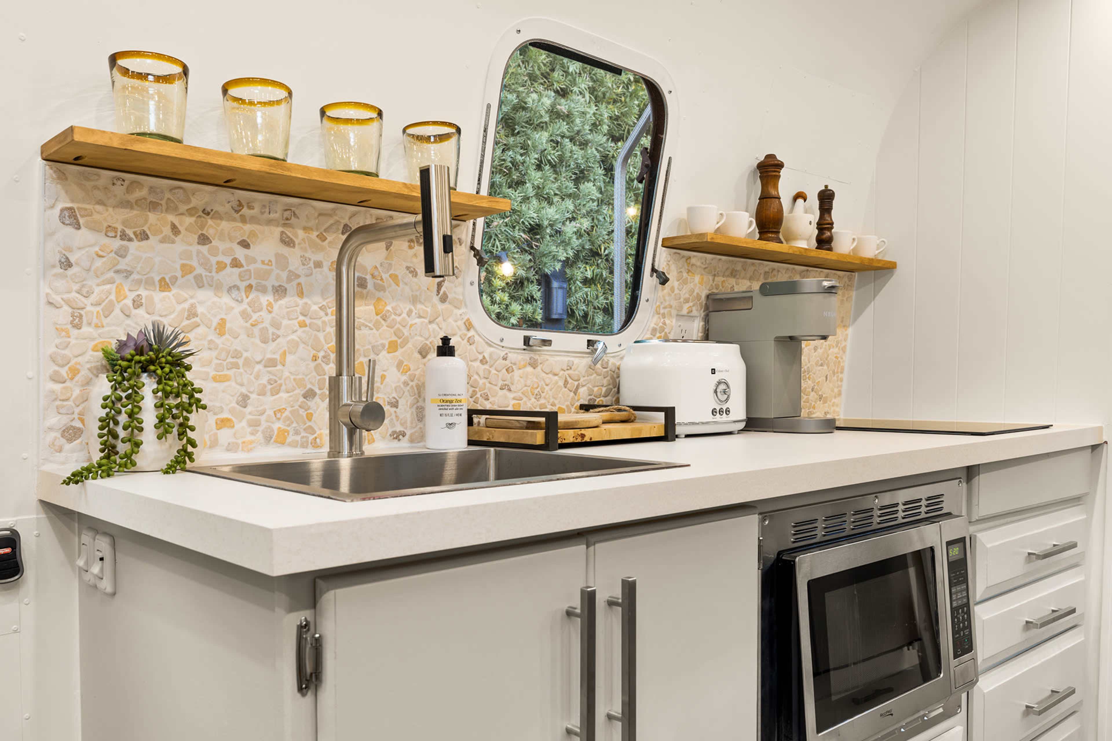 The image shows a modern kitchen area featuring a stainless steel sink, a countertop with decorative backsplash, and wooden shelves displaying various kitchenware.