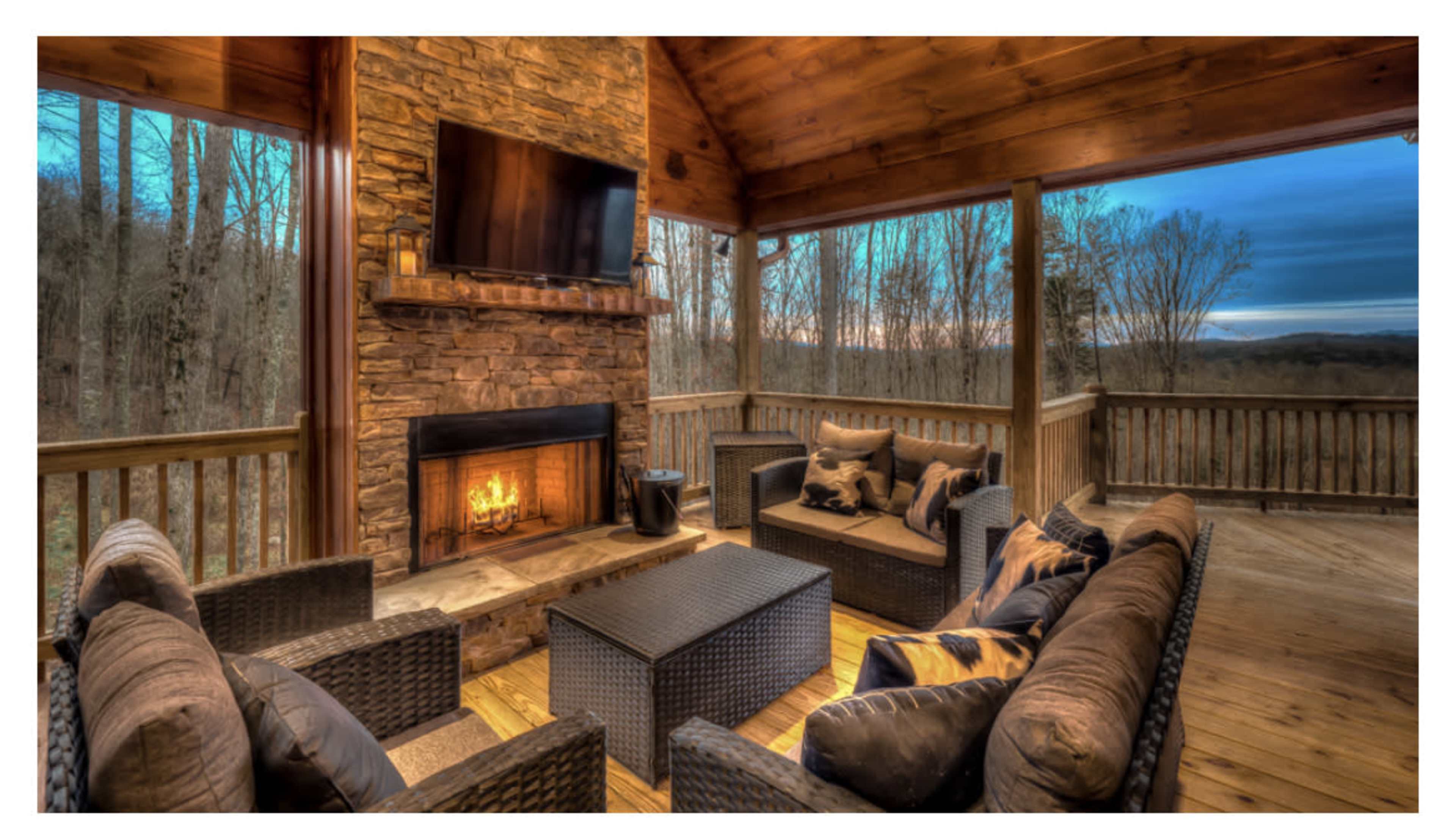 A wooden porch with a stone fireplace, seating area, and a television, surrounded by trees at dusk.