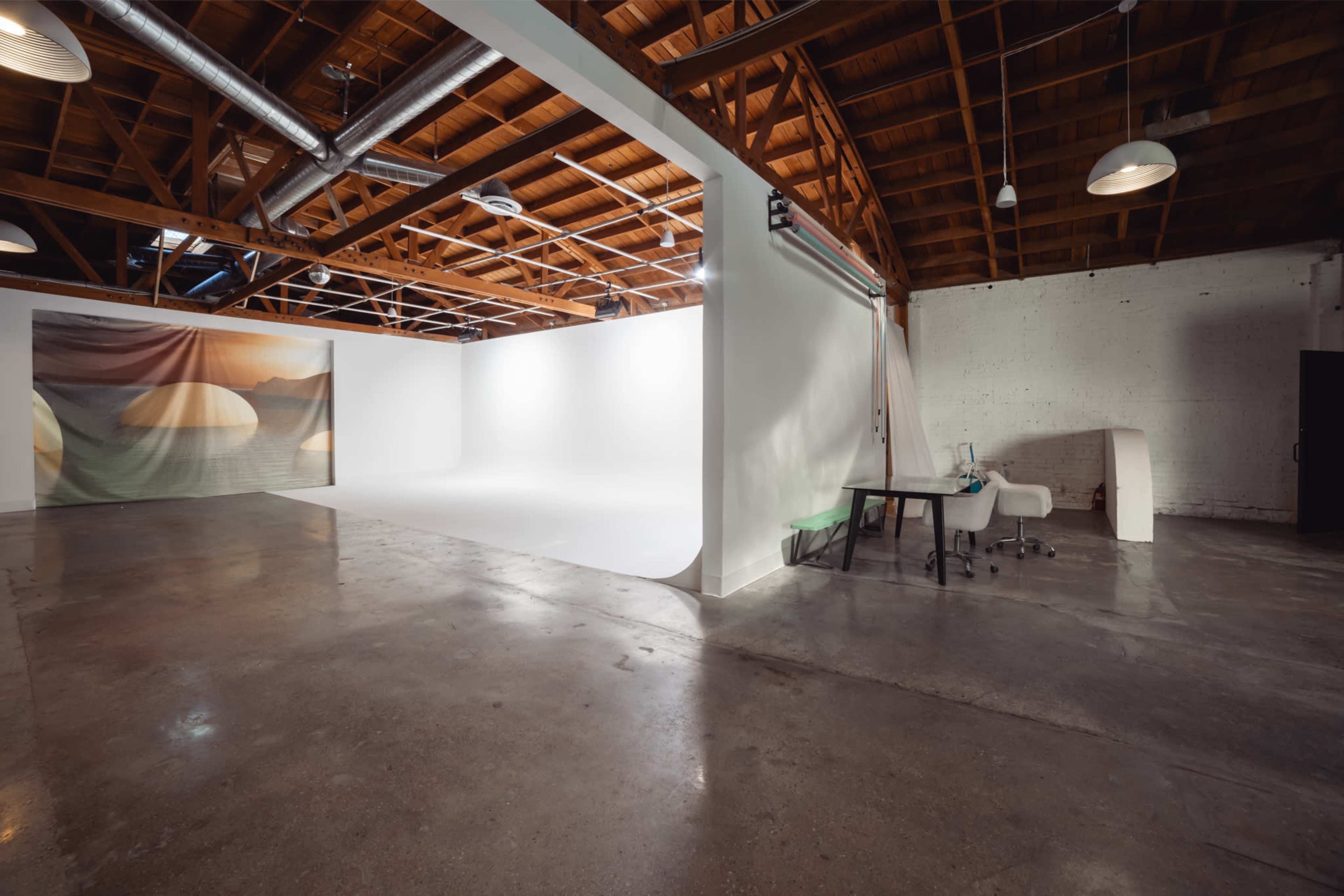 A spacious, well-lit studio with a seamless backdrop, polished concrete floors, and a desk with a chair in the corner.