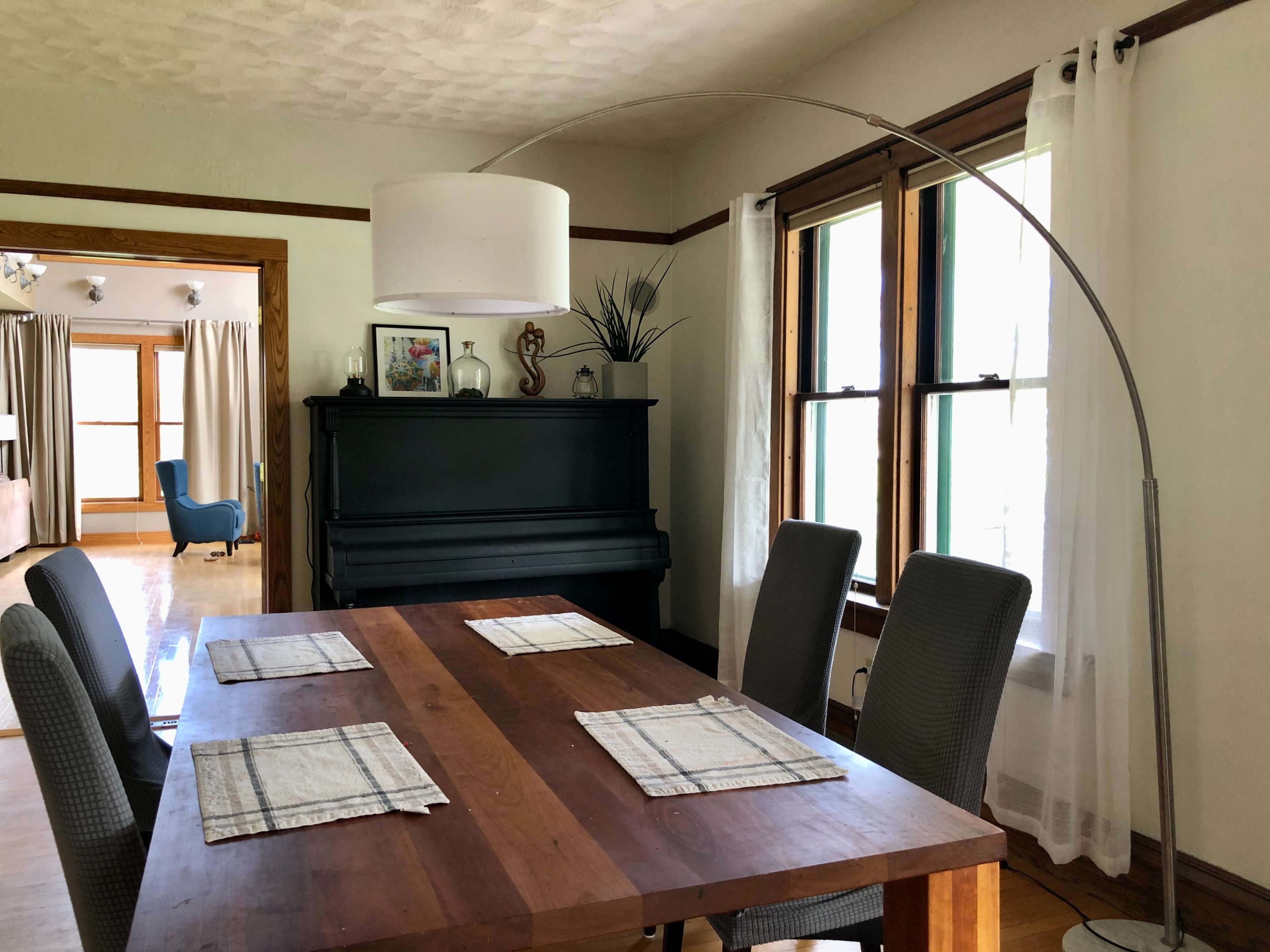 A dining room features a wooden table with placemats, surrounded by six chairs, and an arching floor lamp beside a black piano near two windows.