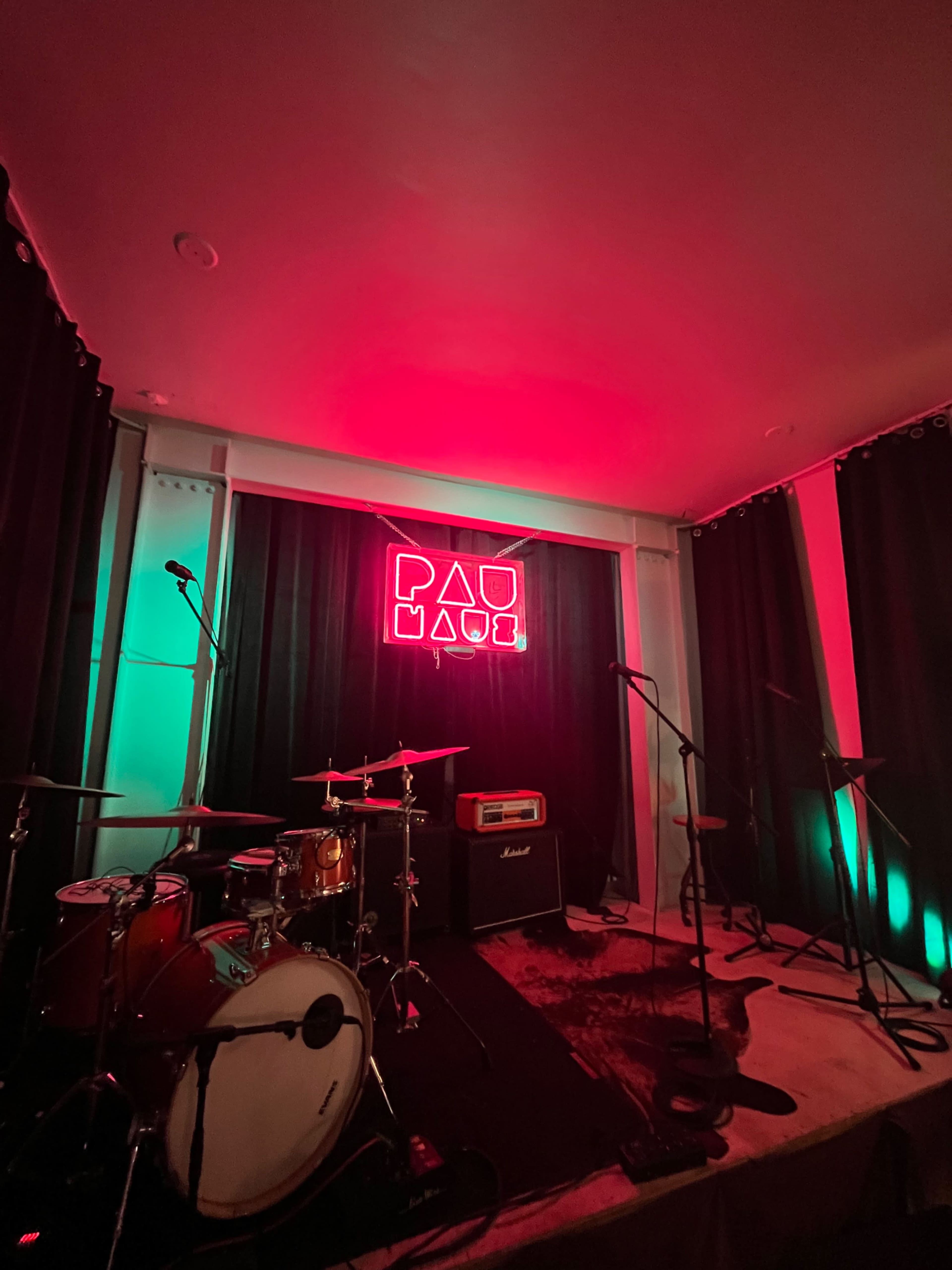 The image shows a small stage with a neon sign that reads "PAU MADE," surrounded by green and red lighting, with musical instruments and microphones positioned in front.