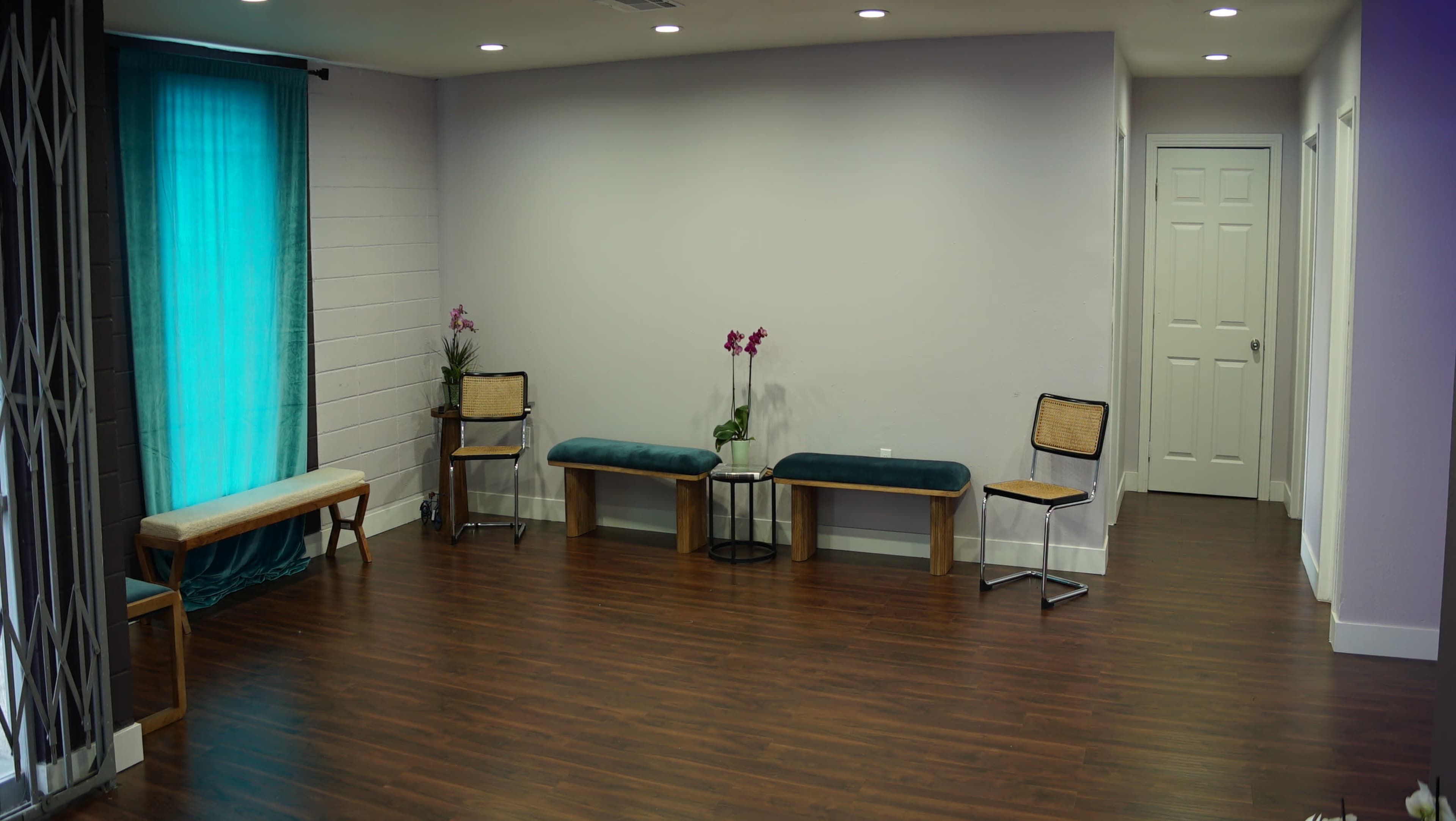 A sparsely furnished room with wooden flooring, featuring two benches, two chairs, and a small table, along with a door and a large window covered by a teal curtain.