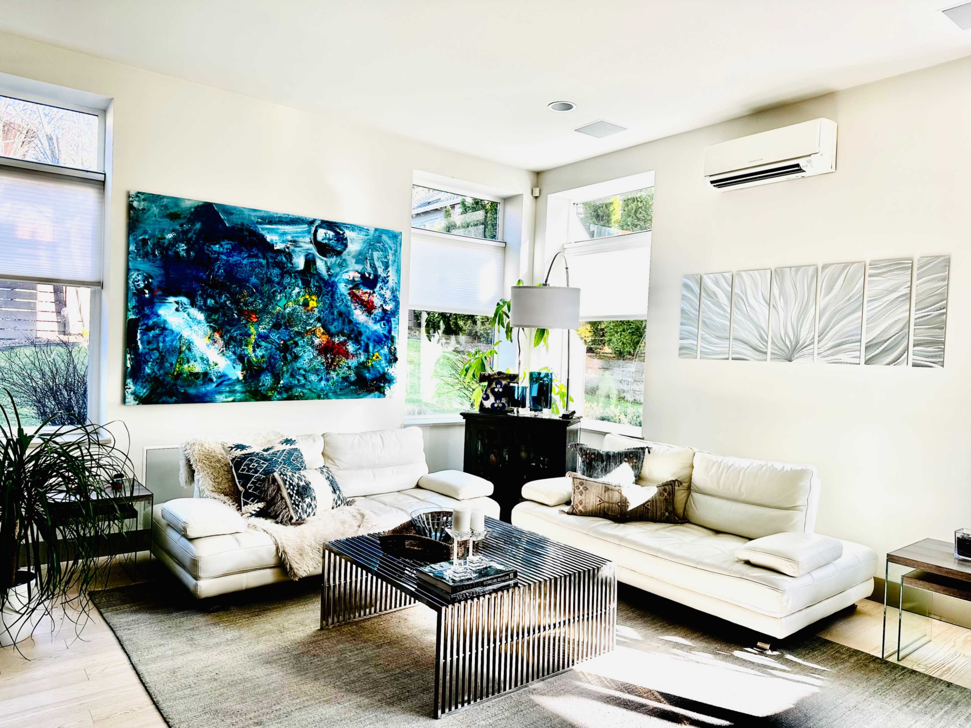 A modern living room with white sofas, a striped coffee table, a large colorful painting on the wall, and plants near the windows.