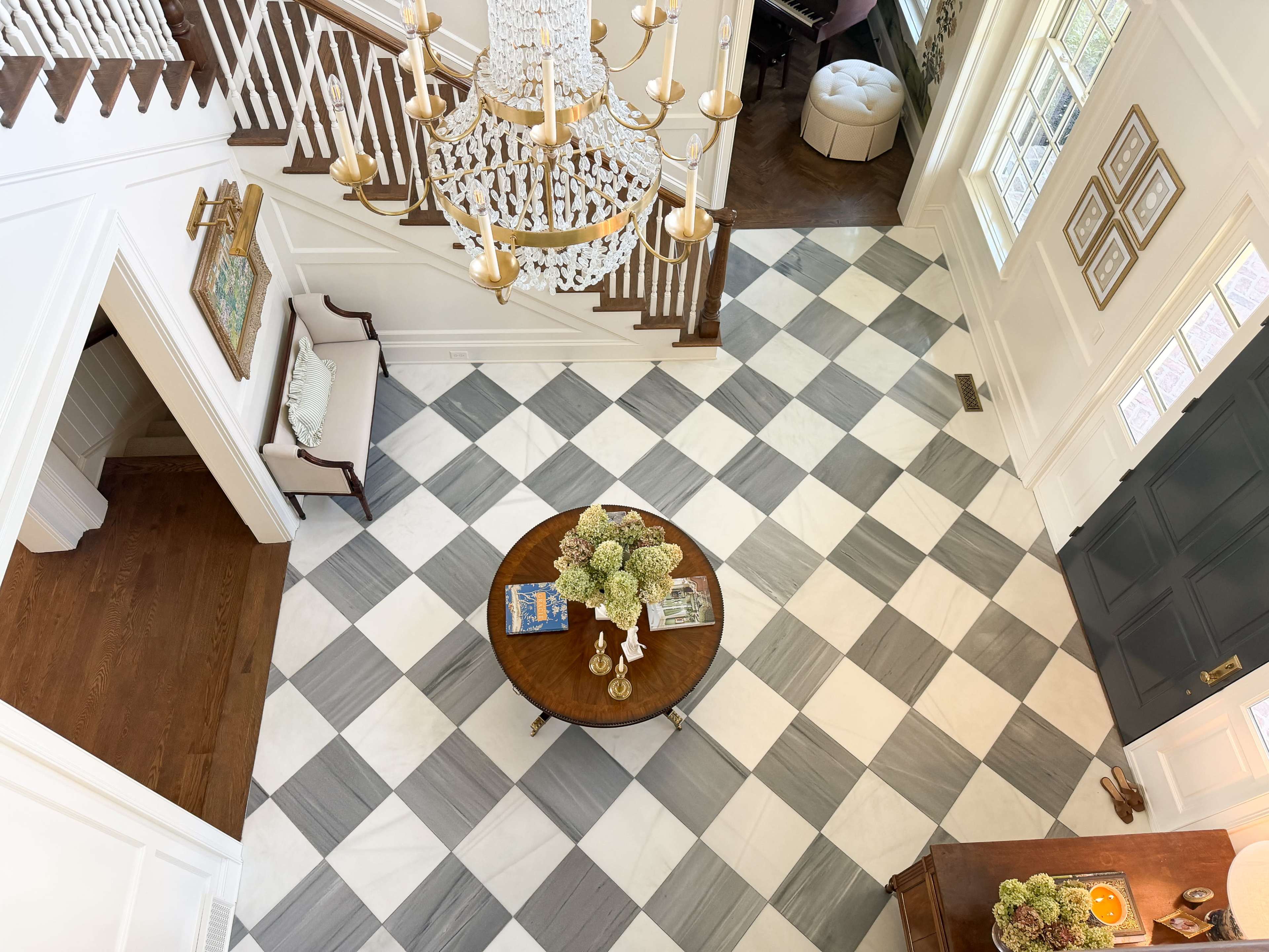 The image shows a spacious entryway with a checkered marble floor, a round wooden table with a floral centerpiece, and a chandelier above.