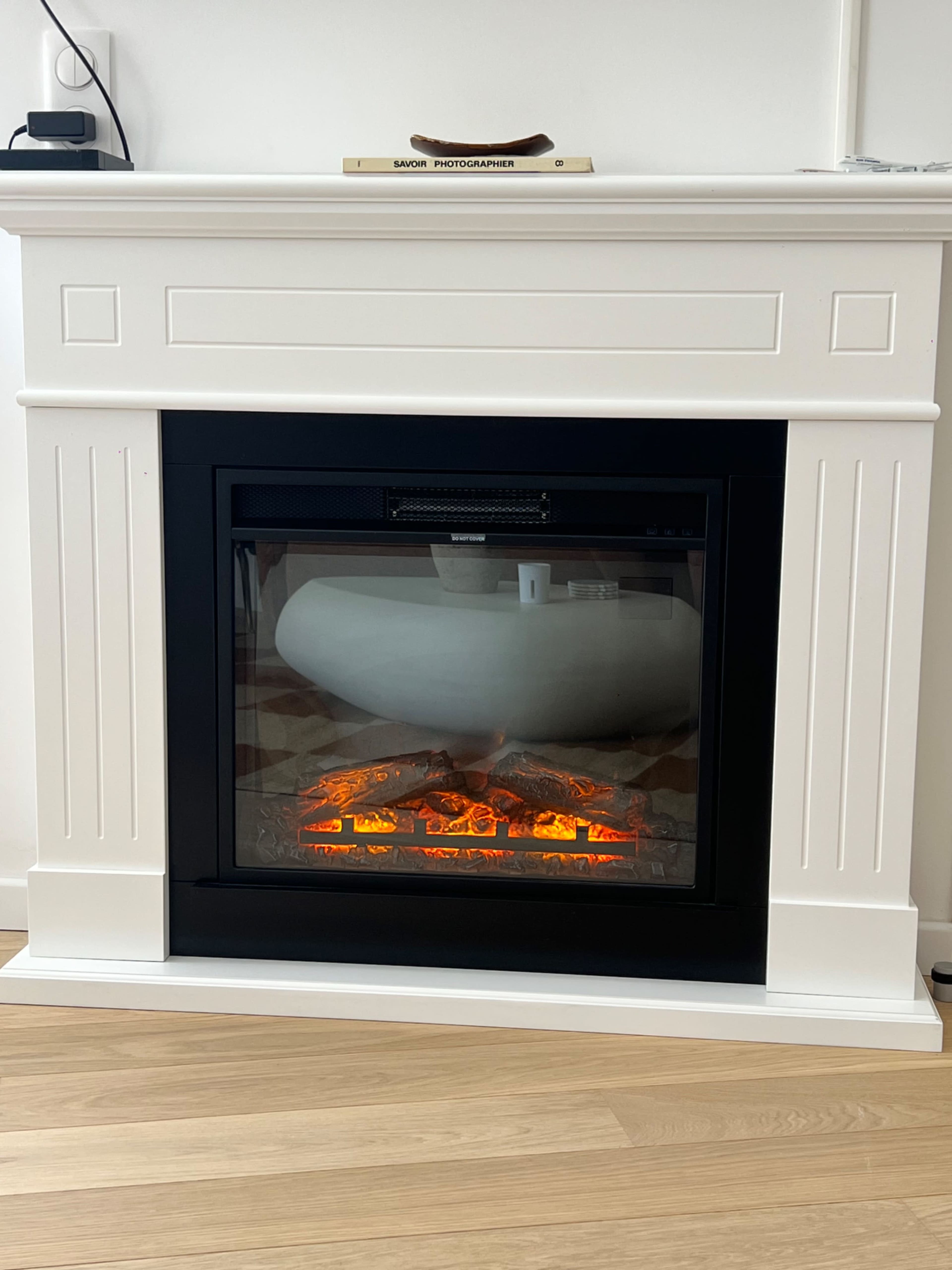 The image shows a modern electric fireplace embedded in a white mantel with a smooth, curved decorative element inside, emitting orange light.
