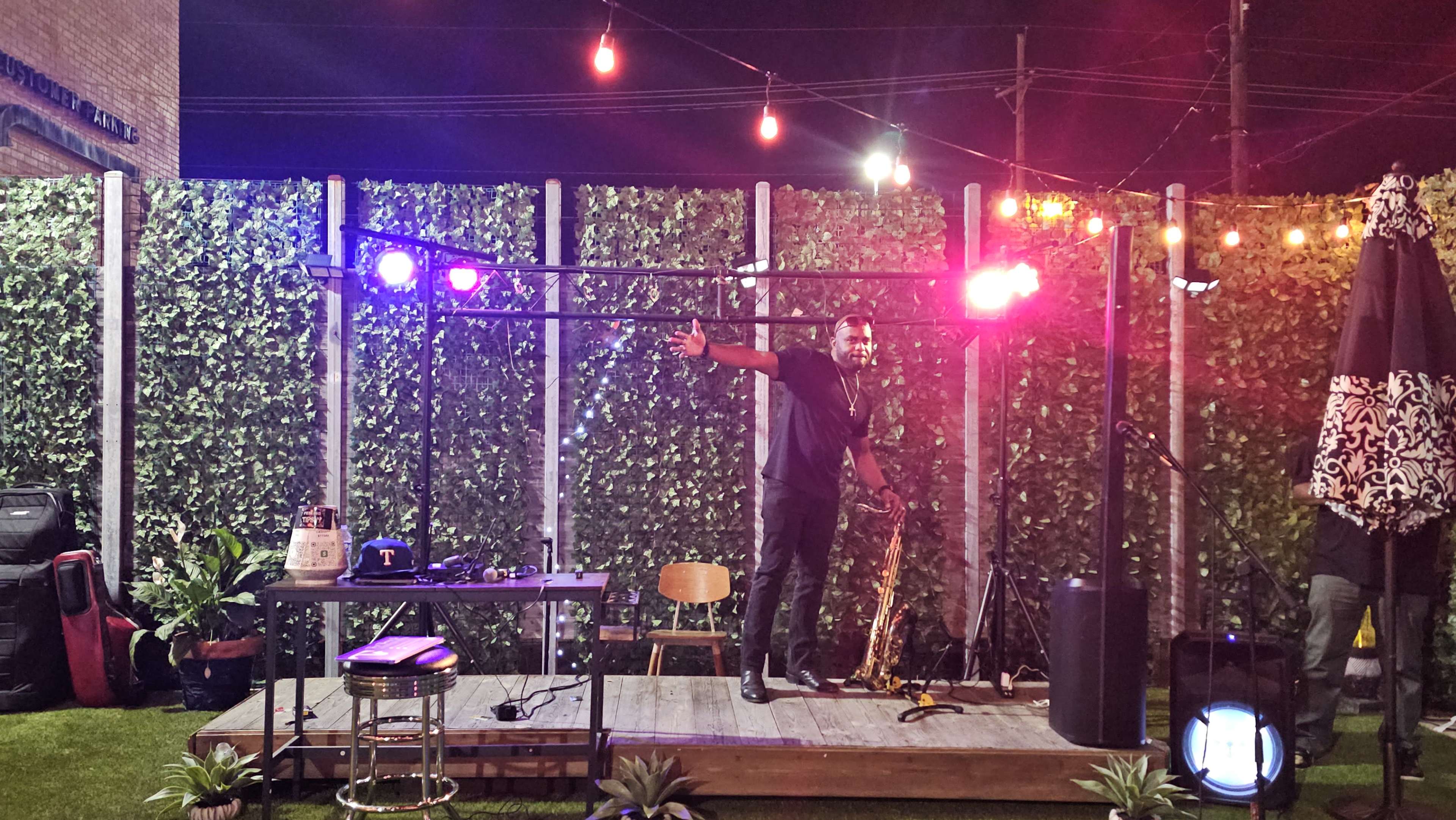 A man stands on a wooden stage under bright lights in a garden setting, surrounded by greenery and various equipment.