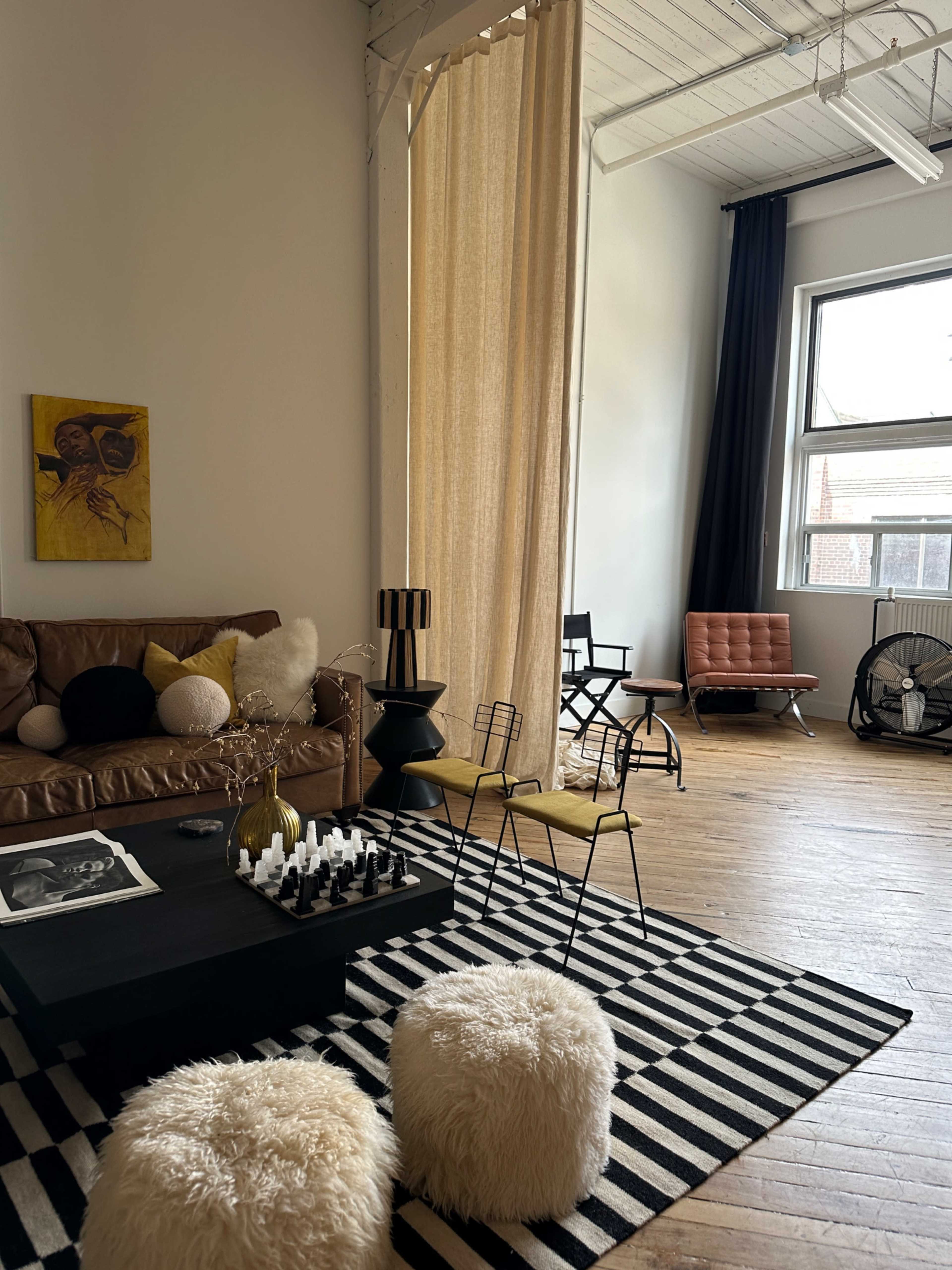The image shows a modern living room with a brown leather sofa, black and white striped rug, a coffee table with decorative items, and a mix of chairs, including a brown armchair and metal stools.