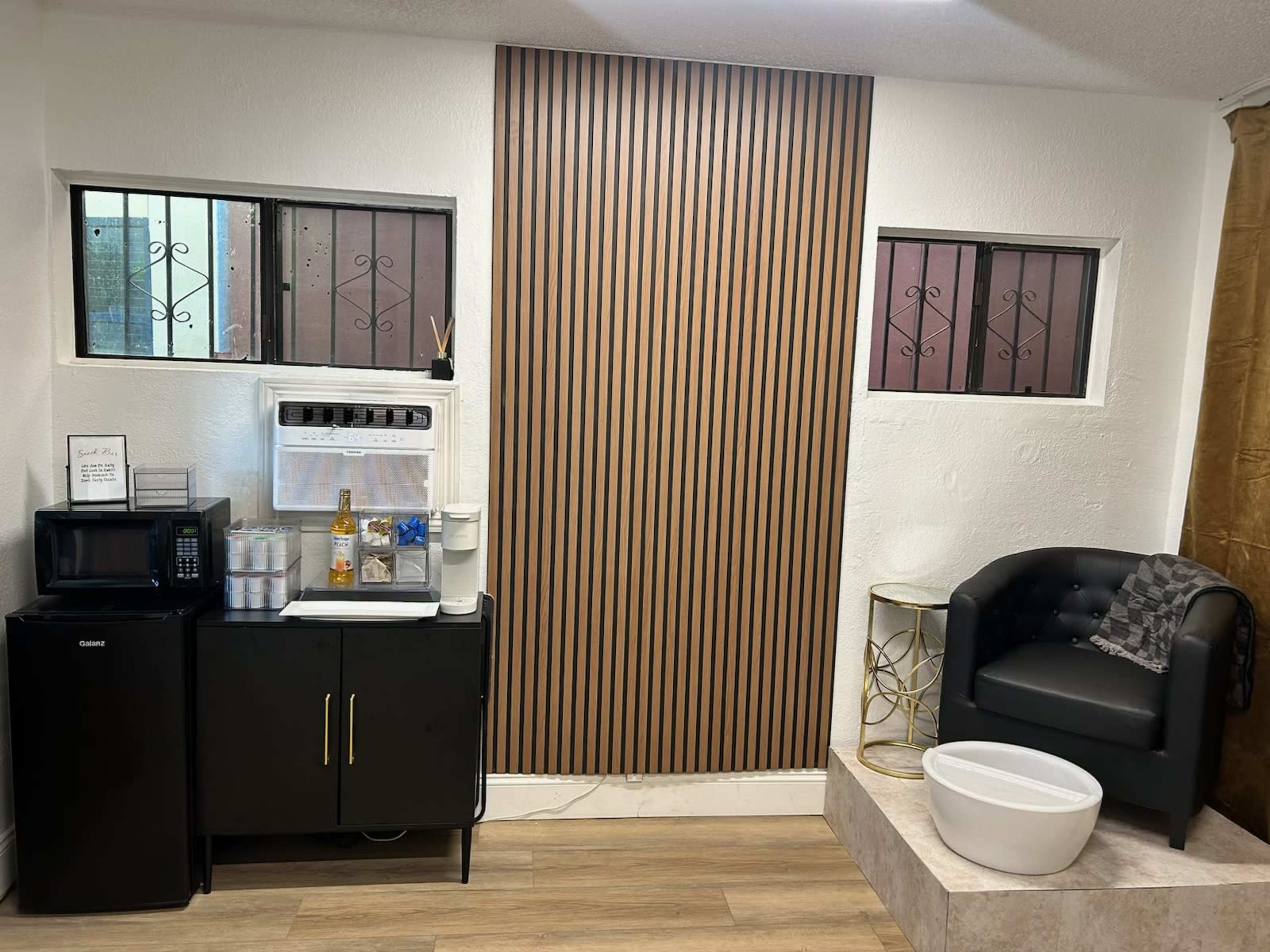 The image shows a small room with a black cabinet, a microwave, a water cooler, a white basin, and a black armchair against a wall with wooden stripes.