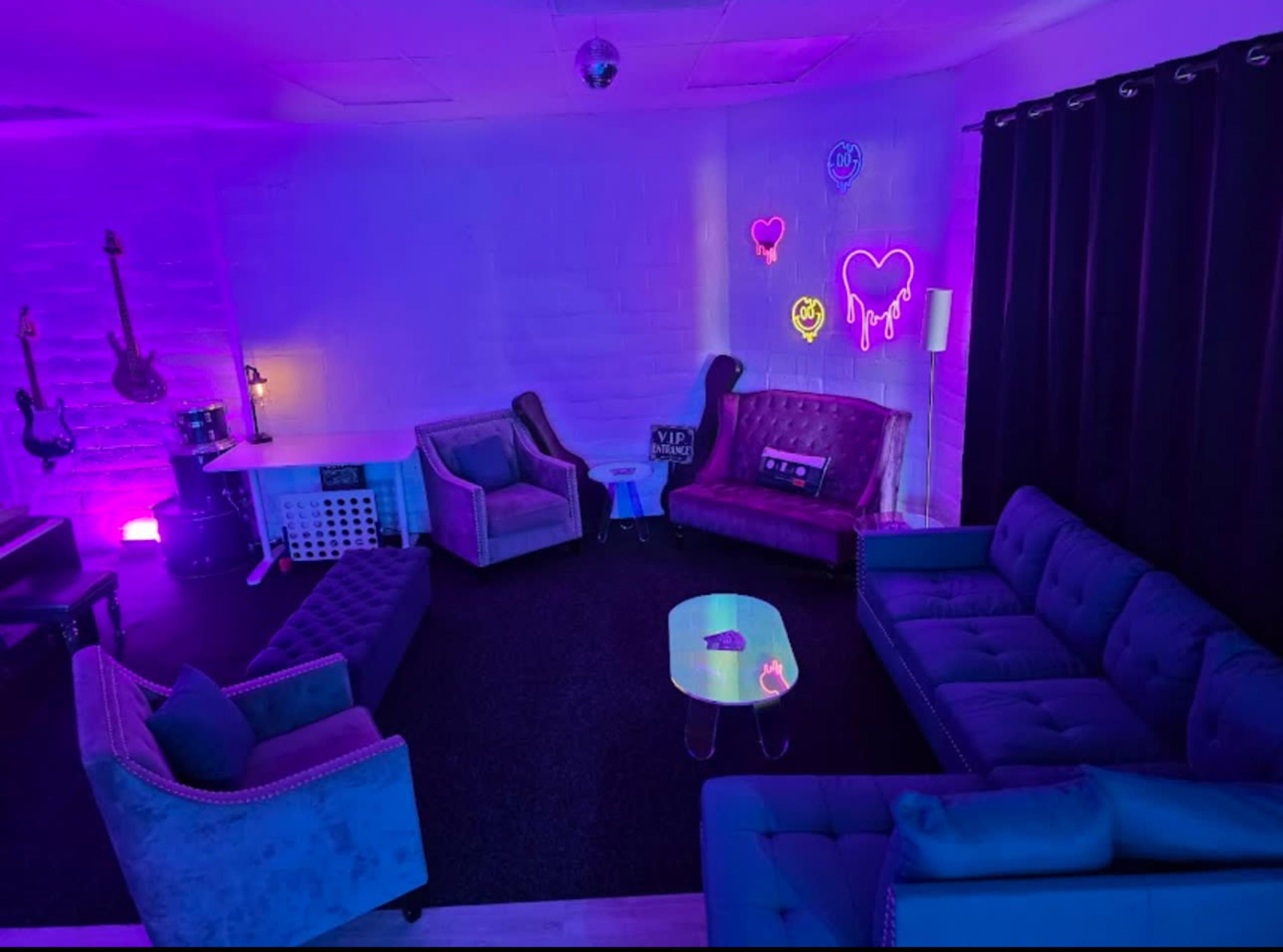 The image shows a dimly lit lounge area with purple and pink neon lights, featuring several sofas arranged around a small table.