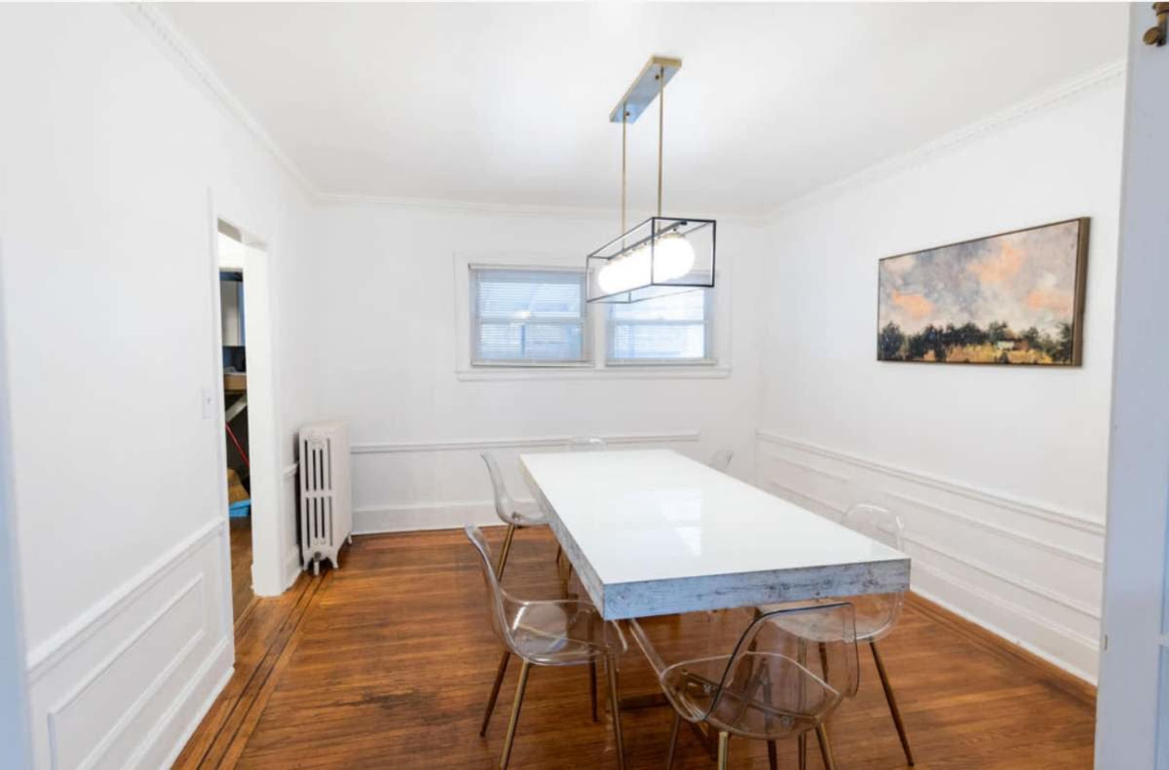 A dining room features a white table with transparent chairs, a pendant light overhead, and a landscape painting on the wall.