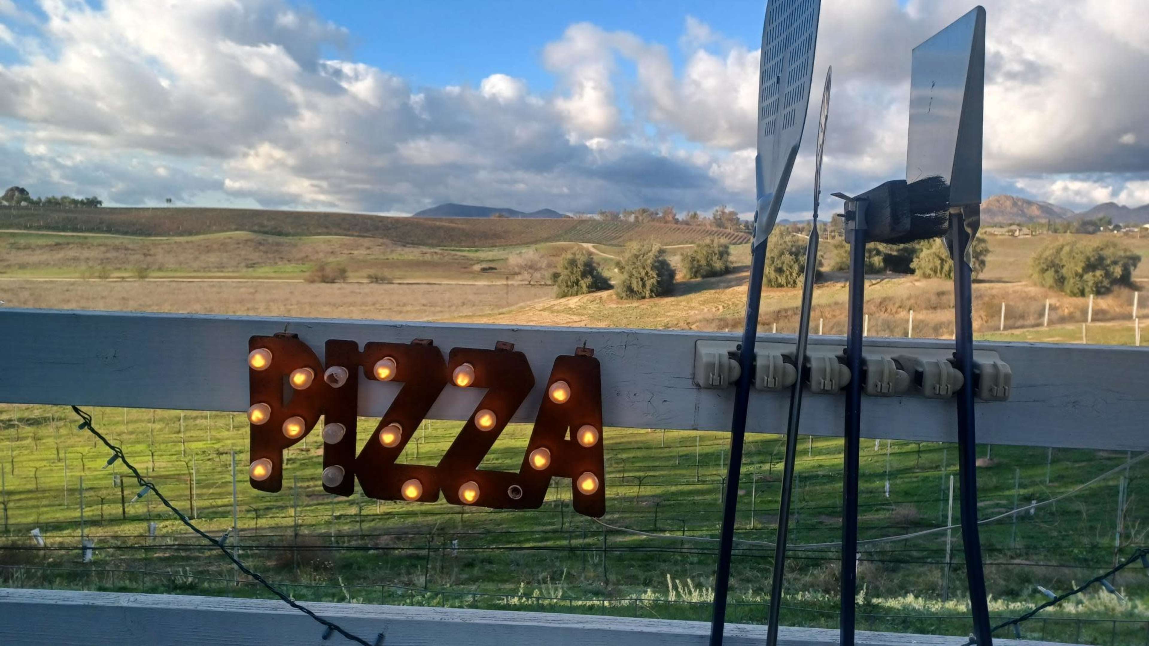 The image shows a rustic sign that reads "PIZZA" with light bulbs, set against a backdrop of rolling green hills and a cloudy sky.