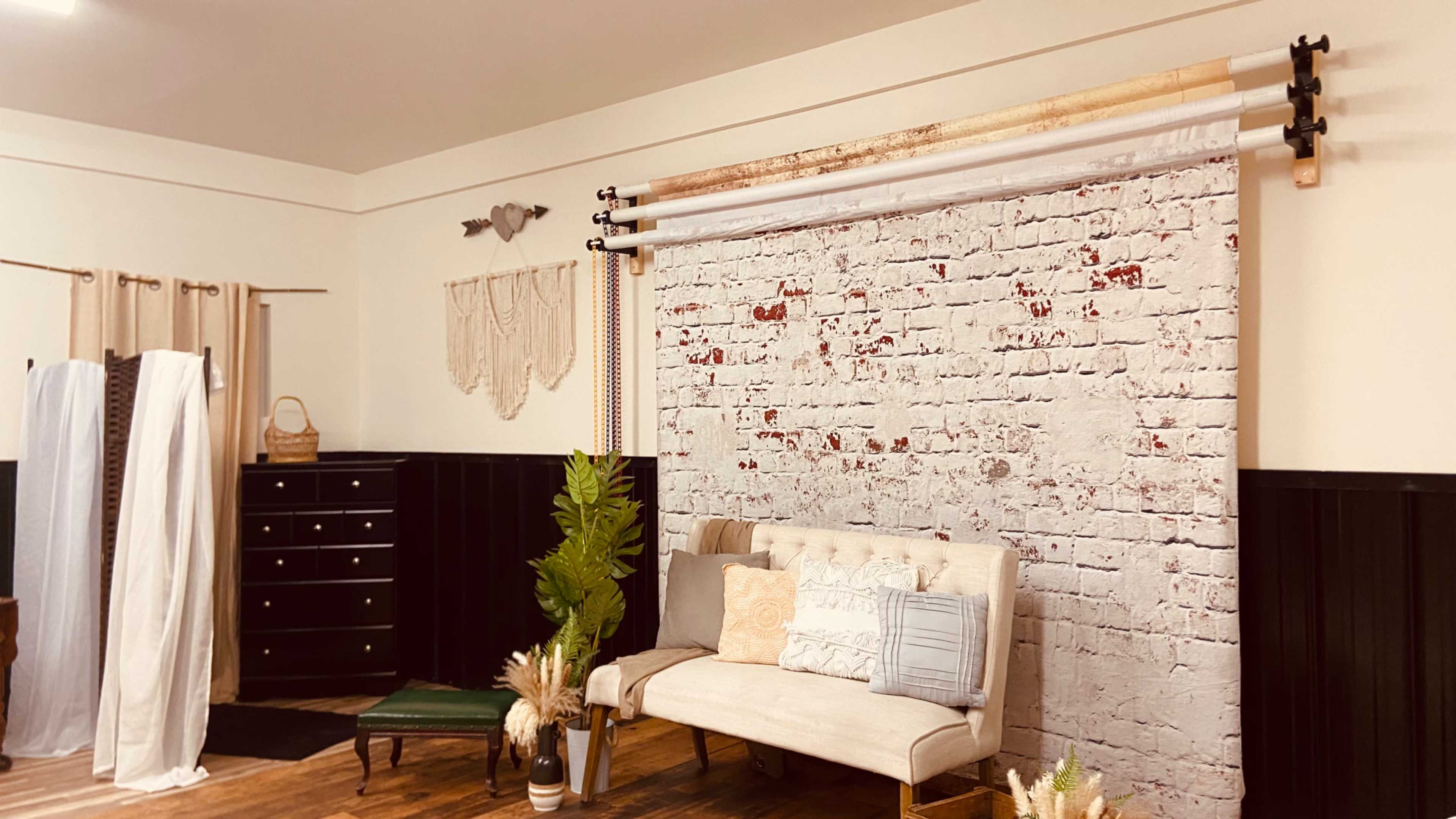 A cozy studio space featuring a white brick backdrop, a light-colored sofa, and various decorative elements including plants and fabric drapes.