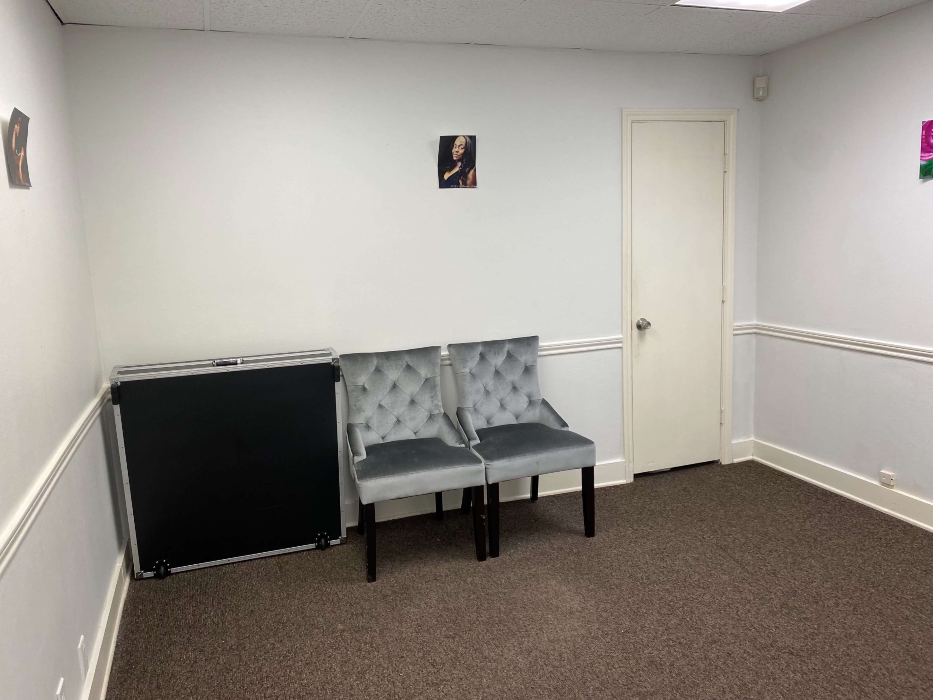 A sparsely furnished room with two gray upholstered chairs against a white wall, a closed door, and a black portable table or storage unit to the side.
