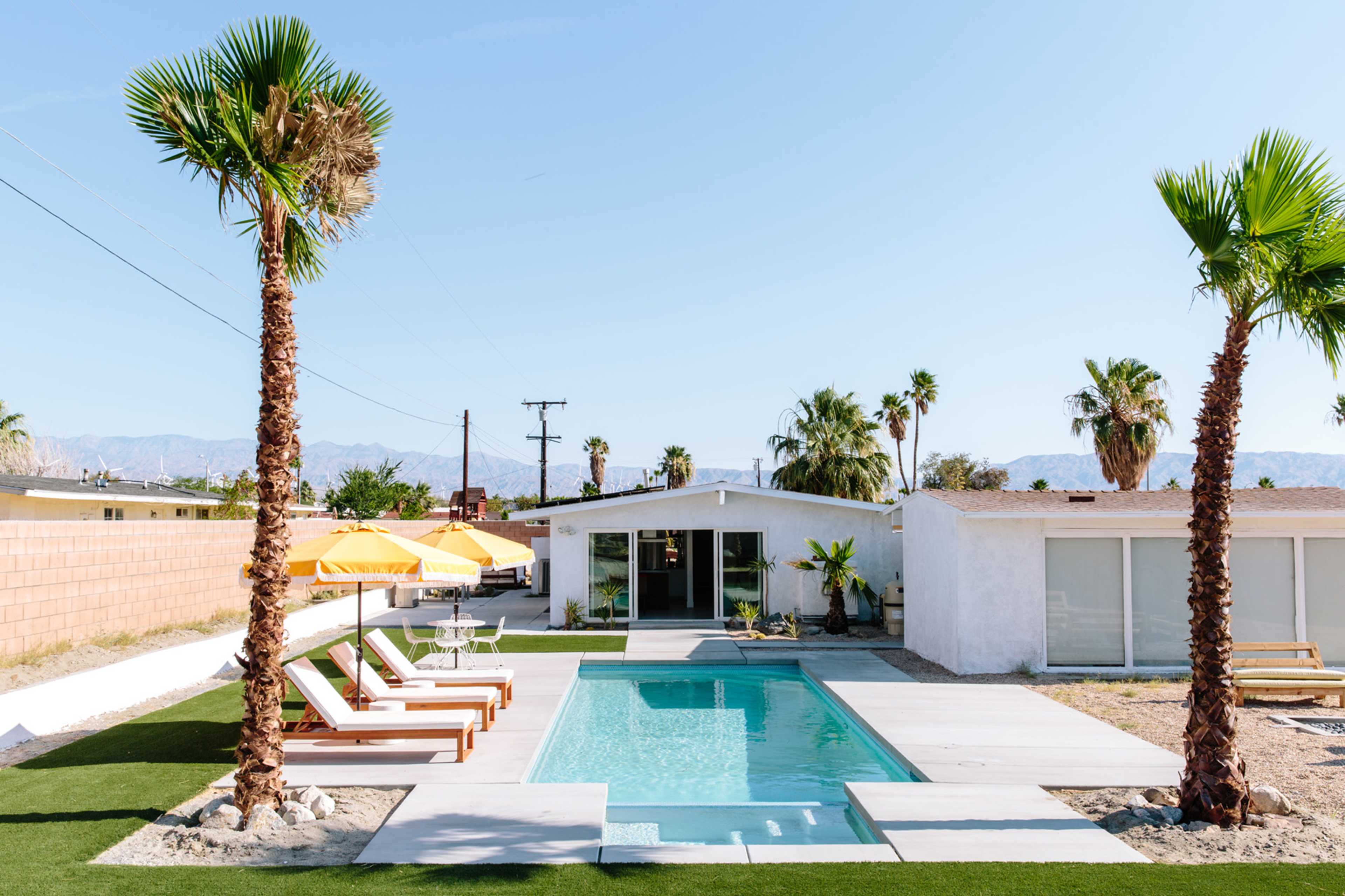 A modern home features a rectangular pool surrounded by lounge chairs and yellow umbrellas, with palm trees flanking the scene against a clear sky.