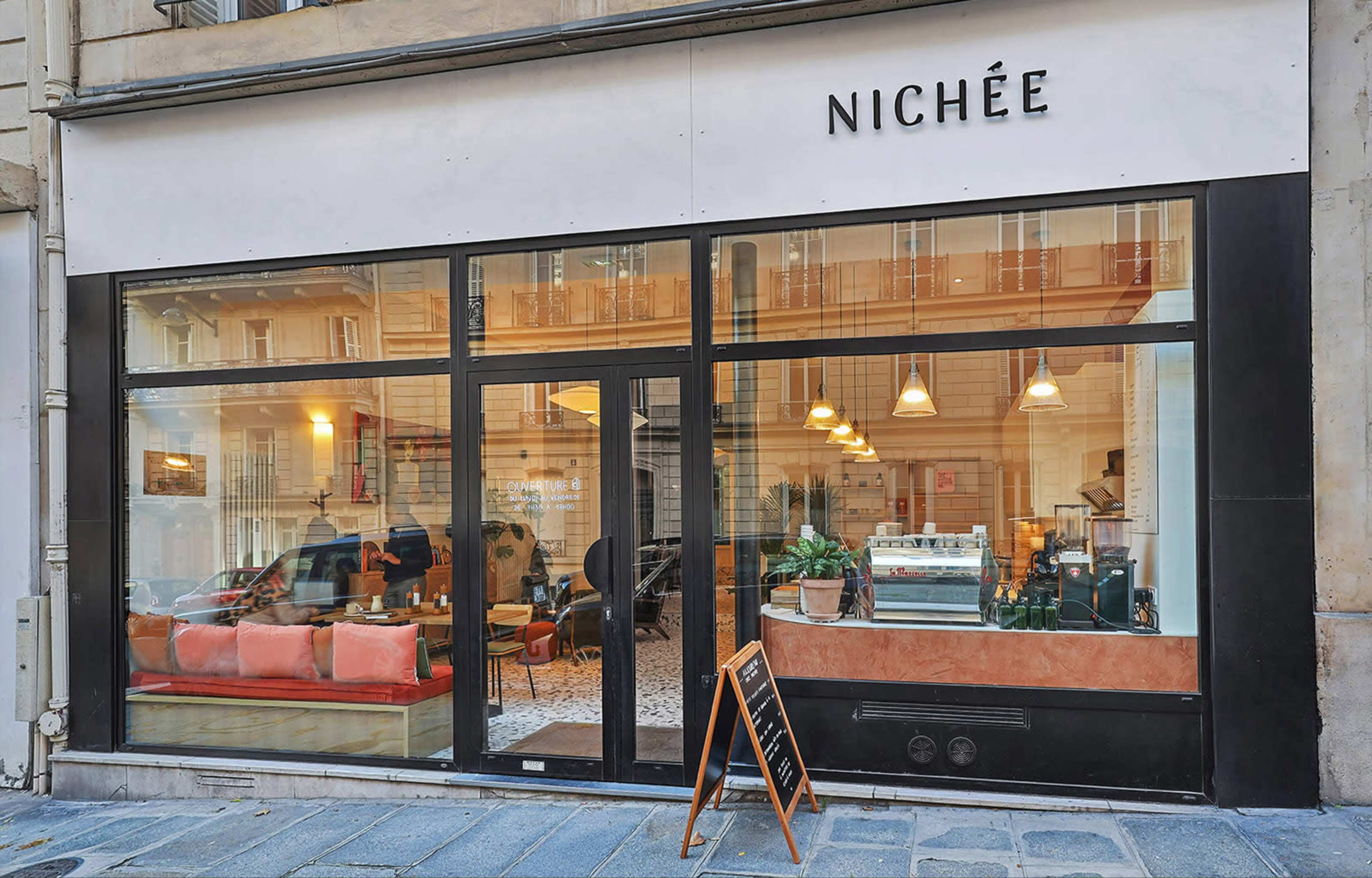 The image shows the exterior of a café named "NICHÉE" with large windows displaying a cozy interior and a sign on an easel outside.