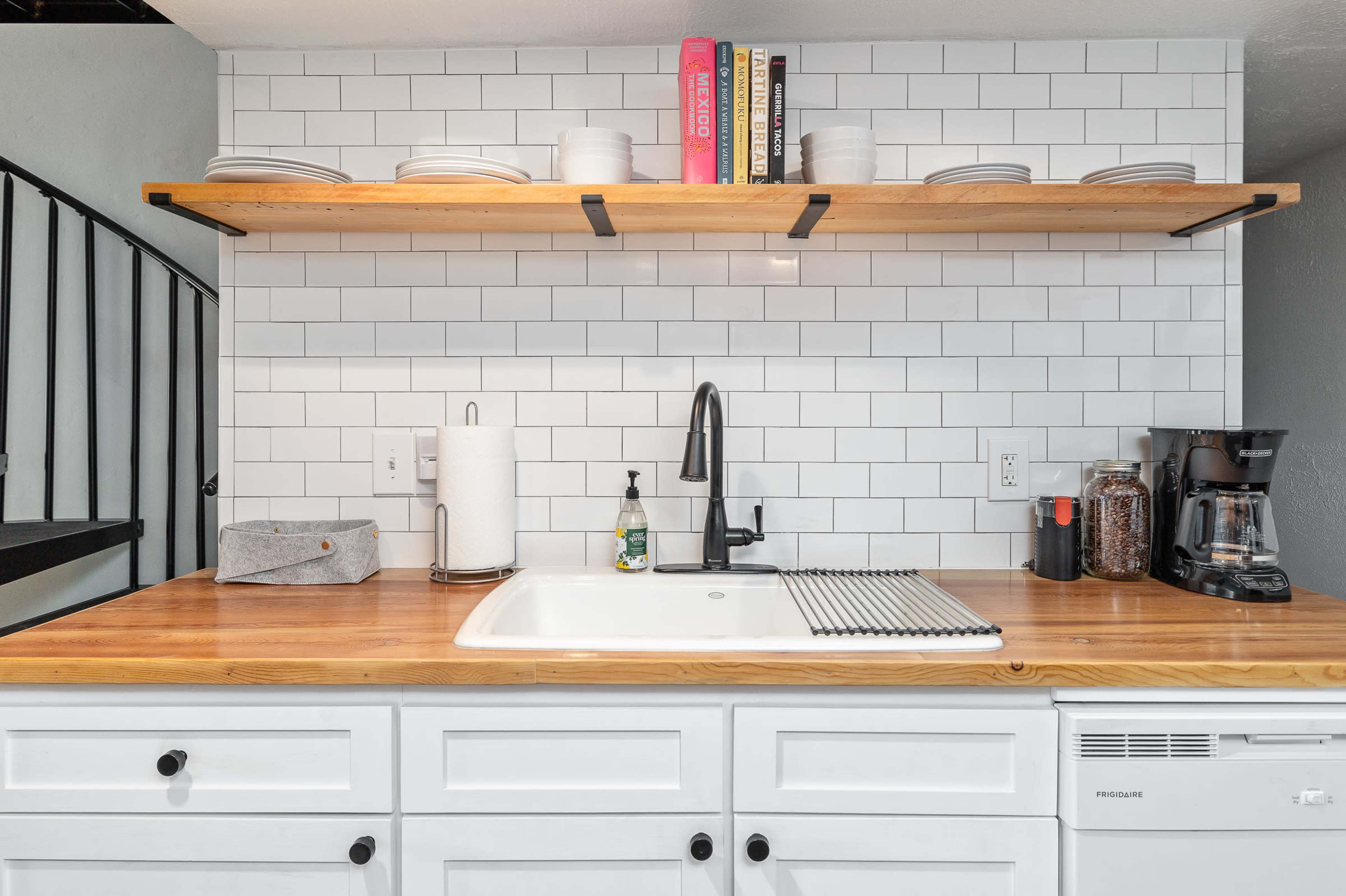 The image shows a modern kitchen with a wooden countertop, a white sink, a wall with white subway tiles, and open shelving displaying plates and books.
