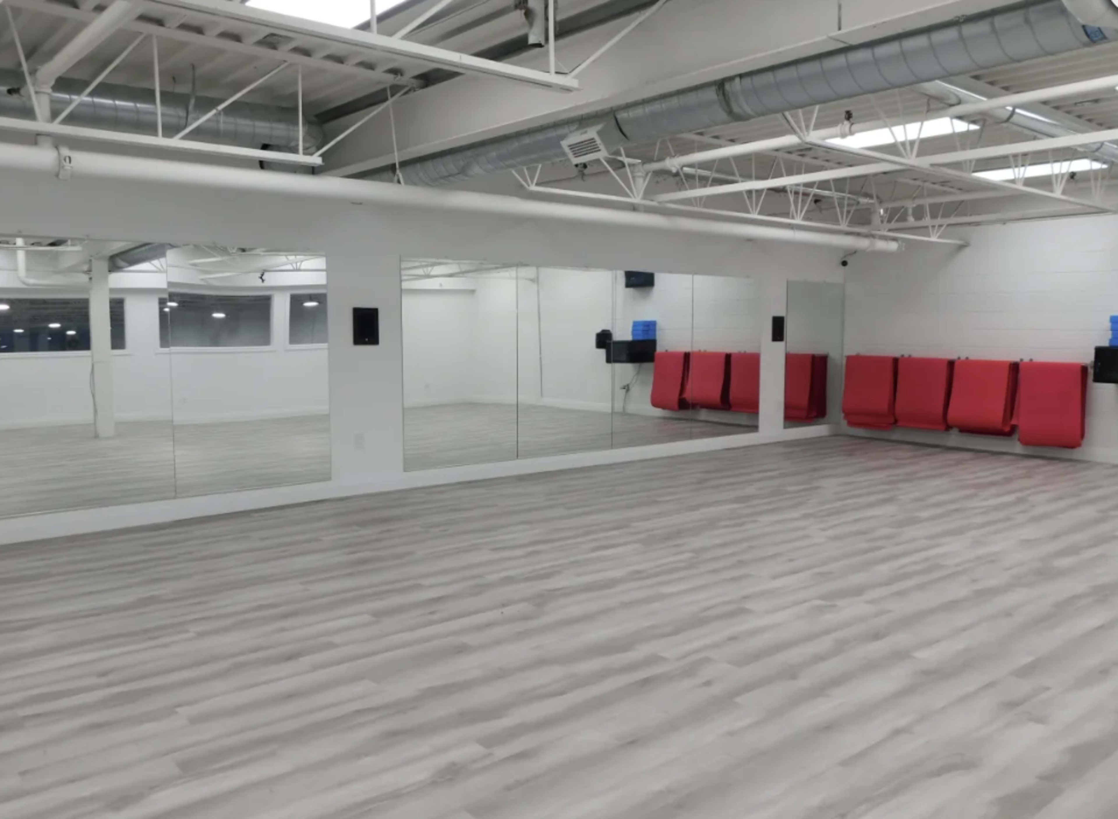 A spacious, empty studio with light-colored wooden flooring, large mirrors on one wall, and red seating against another wall.