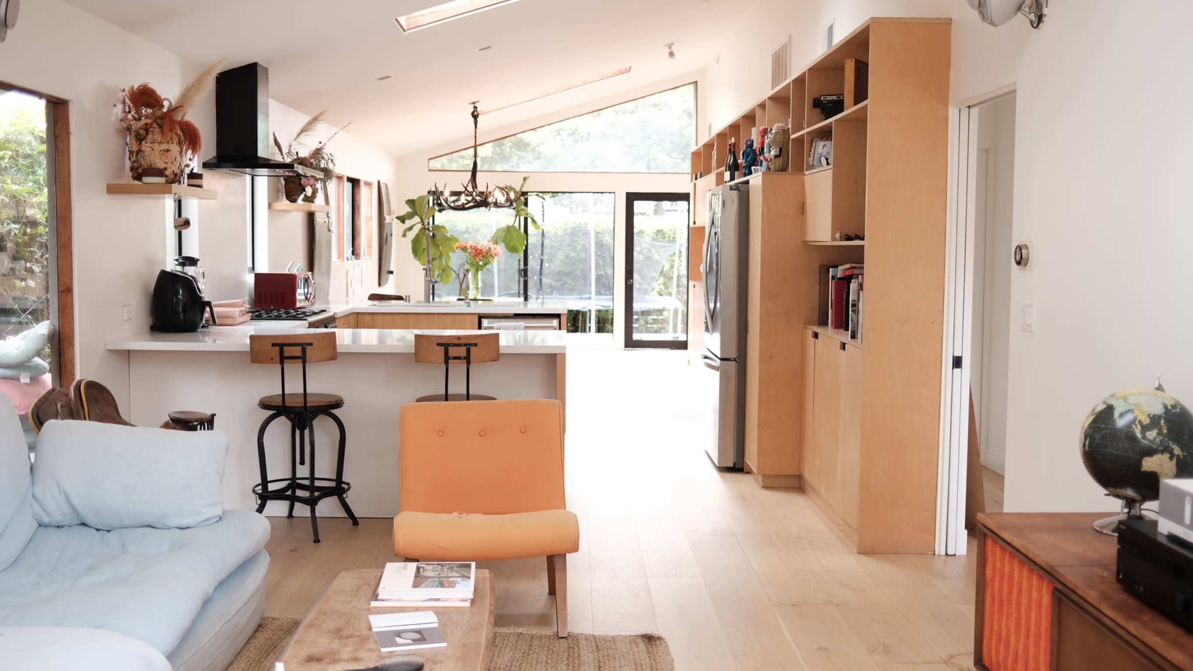 The image shows a bright, modern living space featuring a kitchen with an island, bar stools, and a seating area with a blue couch and orange chair.