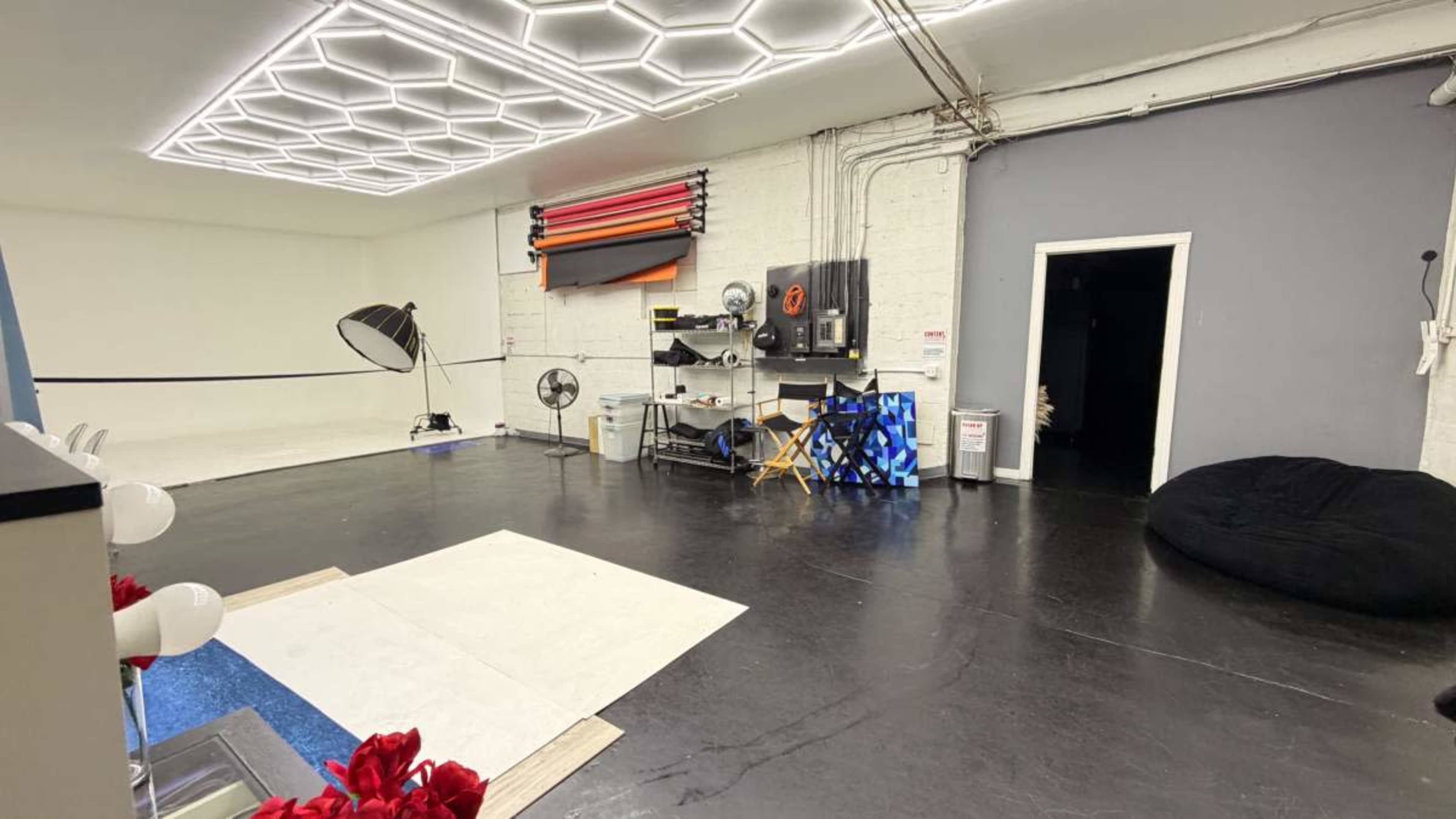 The image shows a professional photography studio with a large seamless backdrop, various lighting equipment, and a seating area.