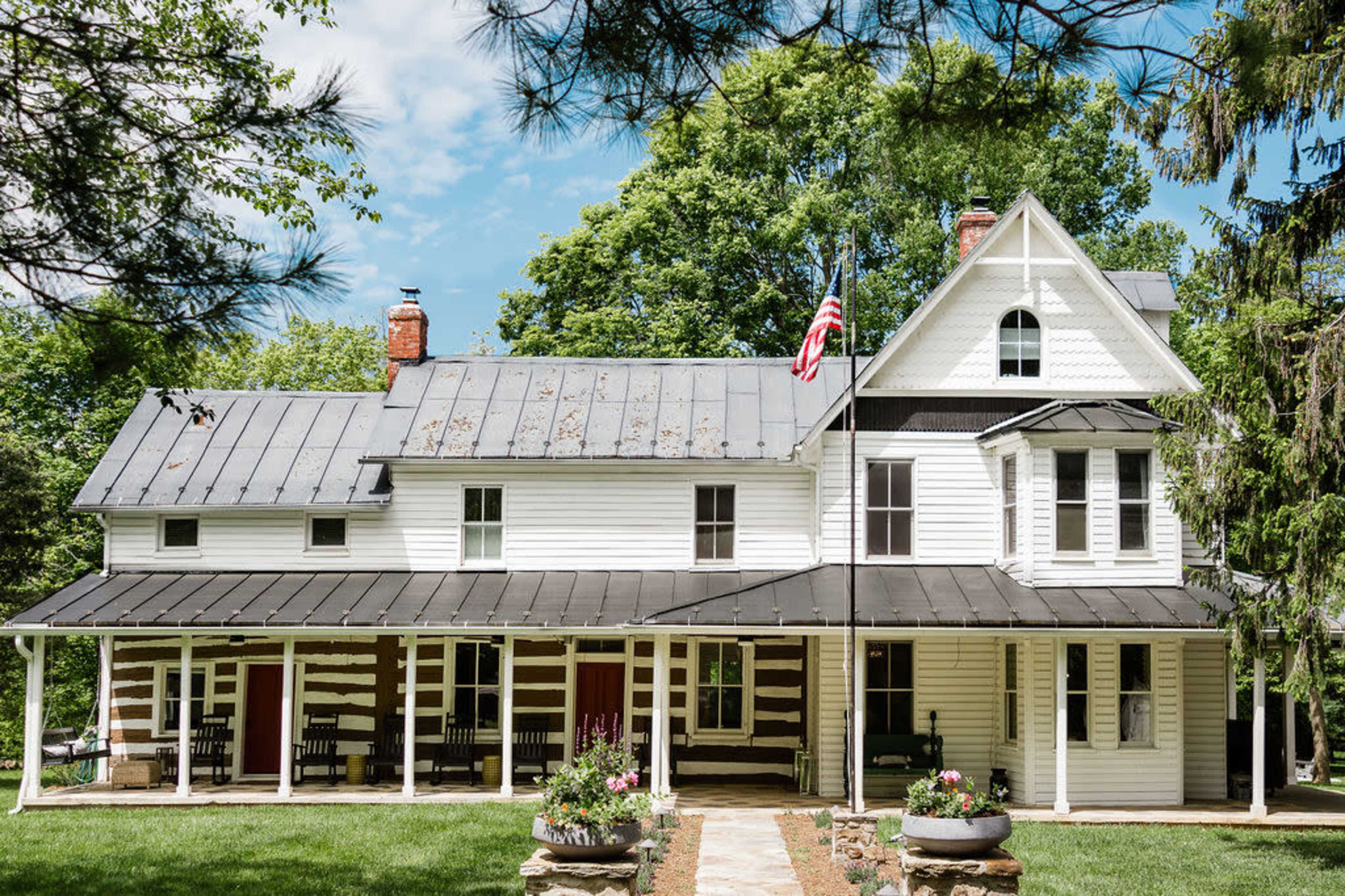 A two-story white farmhouse with a black metal roof, featuring a covered porch and surrounded by trees.