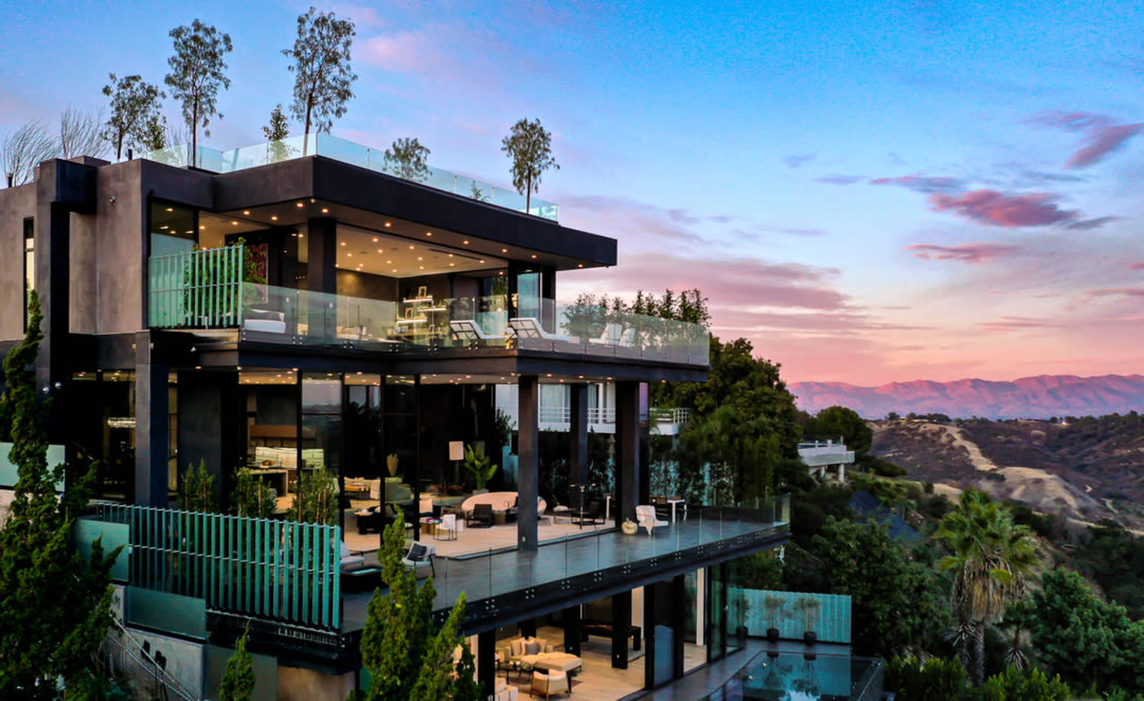 A modern multi-level house with glass balconies is situated on a hillside, surrounded by trees and overlooking distant mountains at sunset.